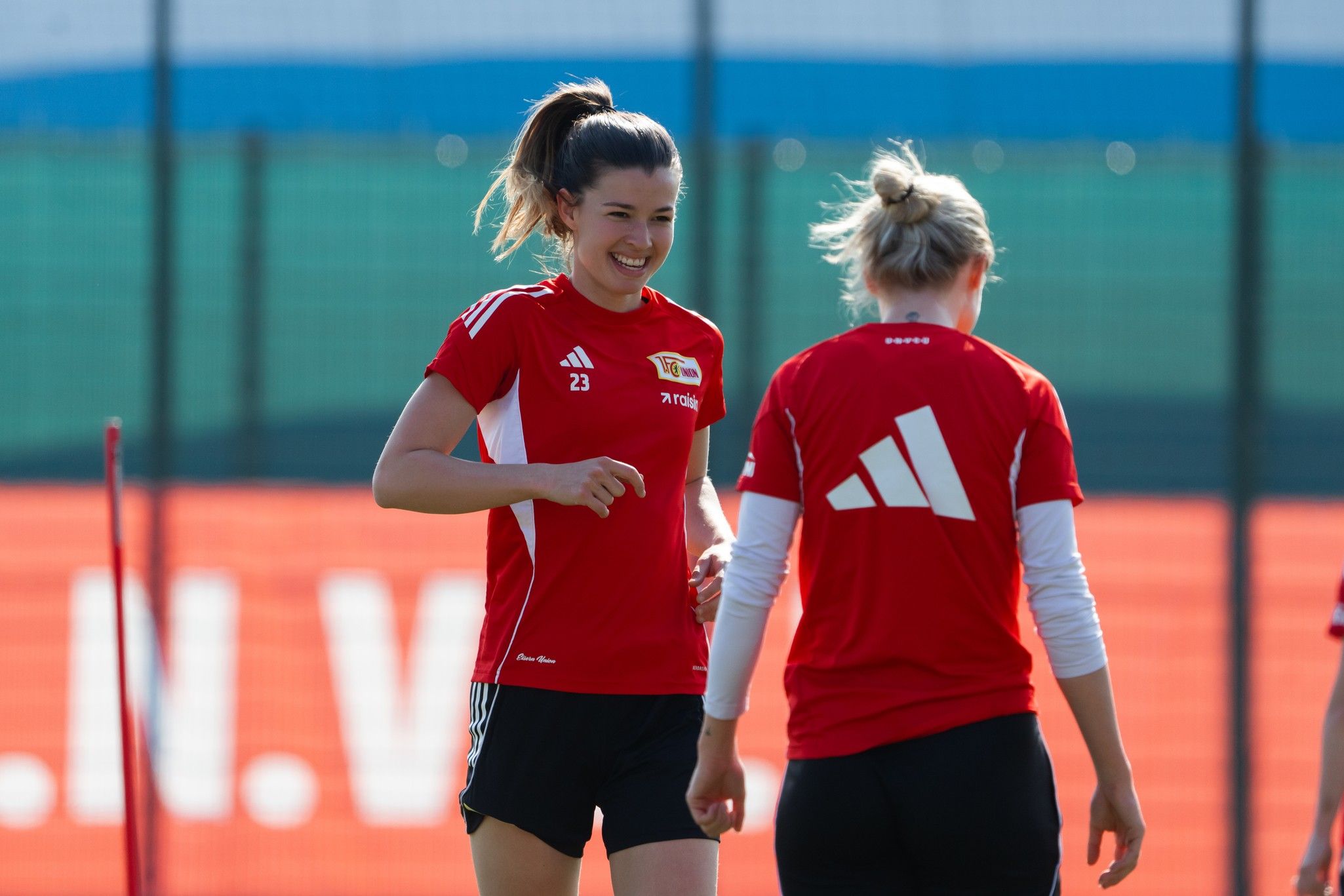Zwei Frauen in roten Trainingsshirts lächeln während des Trainings auf dem Fußballplatz.