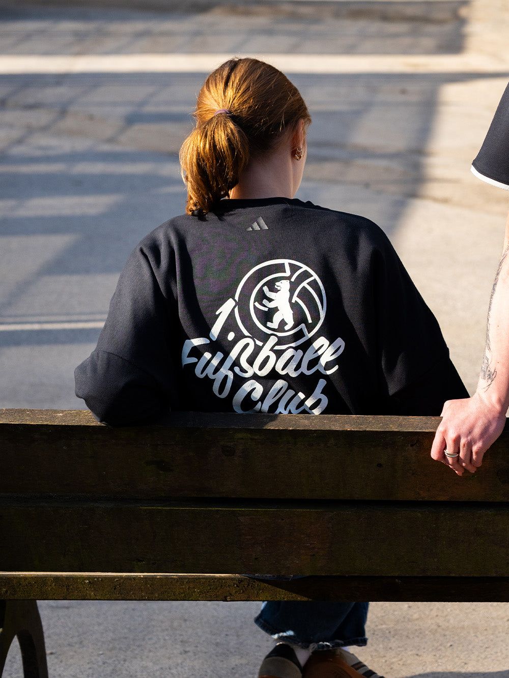 Eine Person mit roten Haaren sitzt auf einer Bank und trägt ein schwarzes Sweatshirt mit der Aufschrift "Lifespace Club".