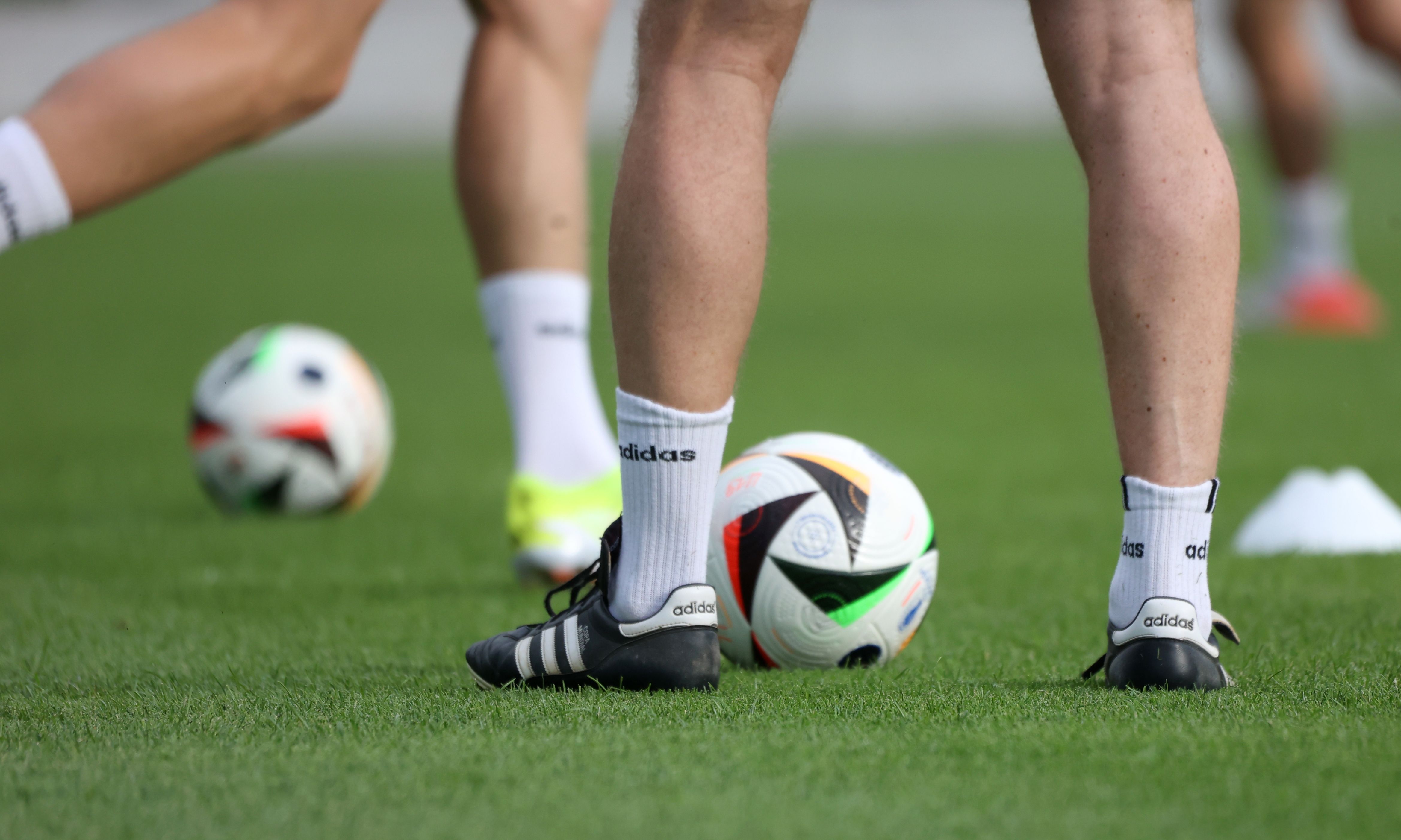 Füße von Spielern in Fußballschuhen auf grünem Rasen, umgeben von mehreren Fußbällen.