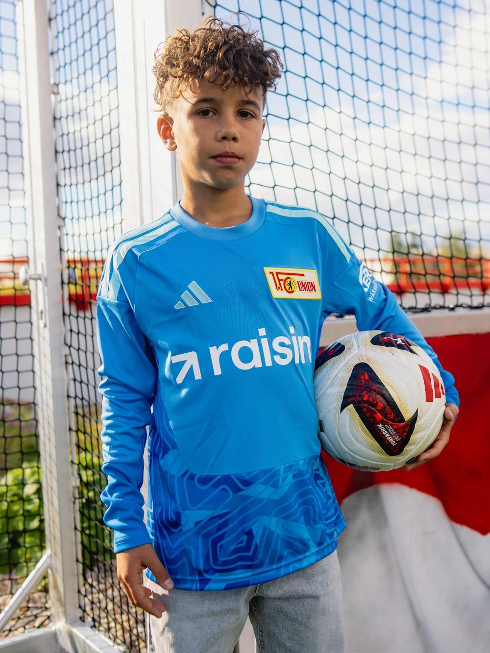 Ein Junge steht vor einem Fußballtor, trägt ein blaues Trikot und hält einen Fußball in der Hand.