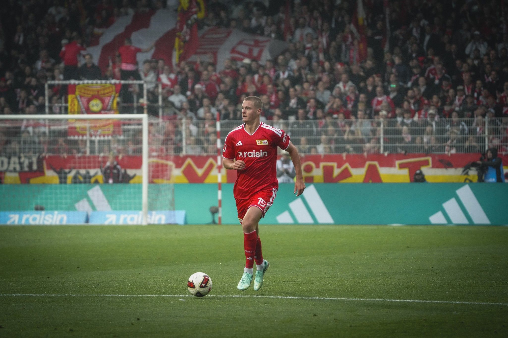Ein Fußballspieler in rotem Trikot läuft auf dem Spielfeld, im Hintergrund Fans mit Fahnen und Banner.
