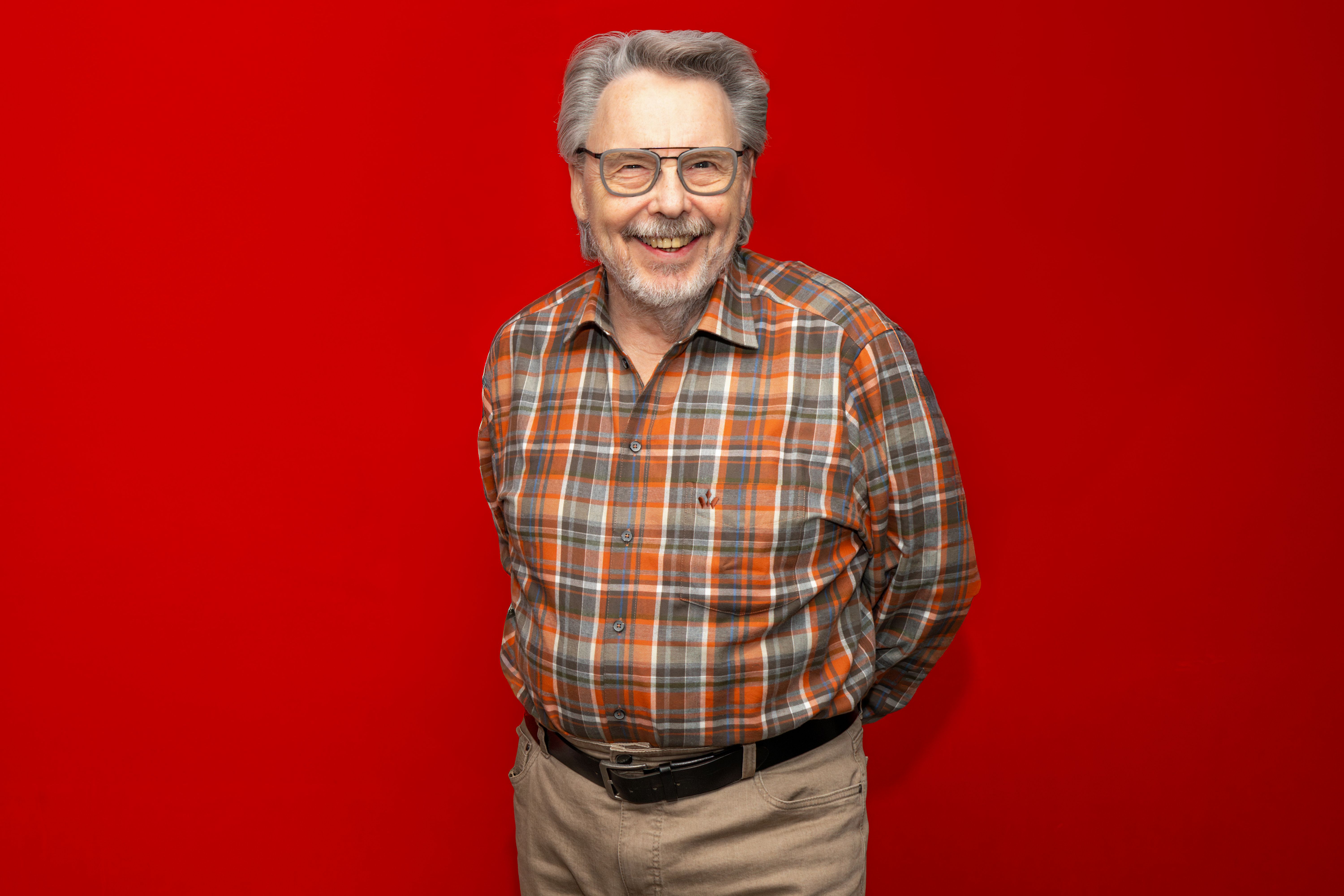 Ein lächelnder älterer Mann mit Brille trägt ein kariertes Hemd vor einem roten Hintergrund.
