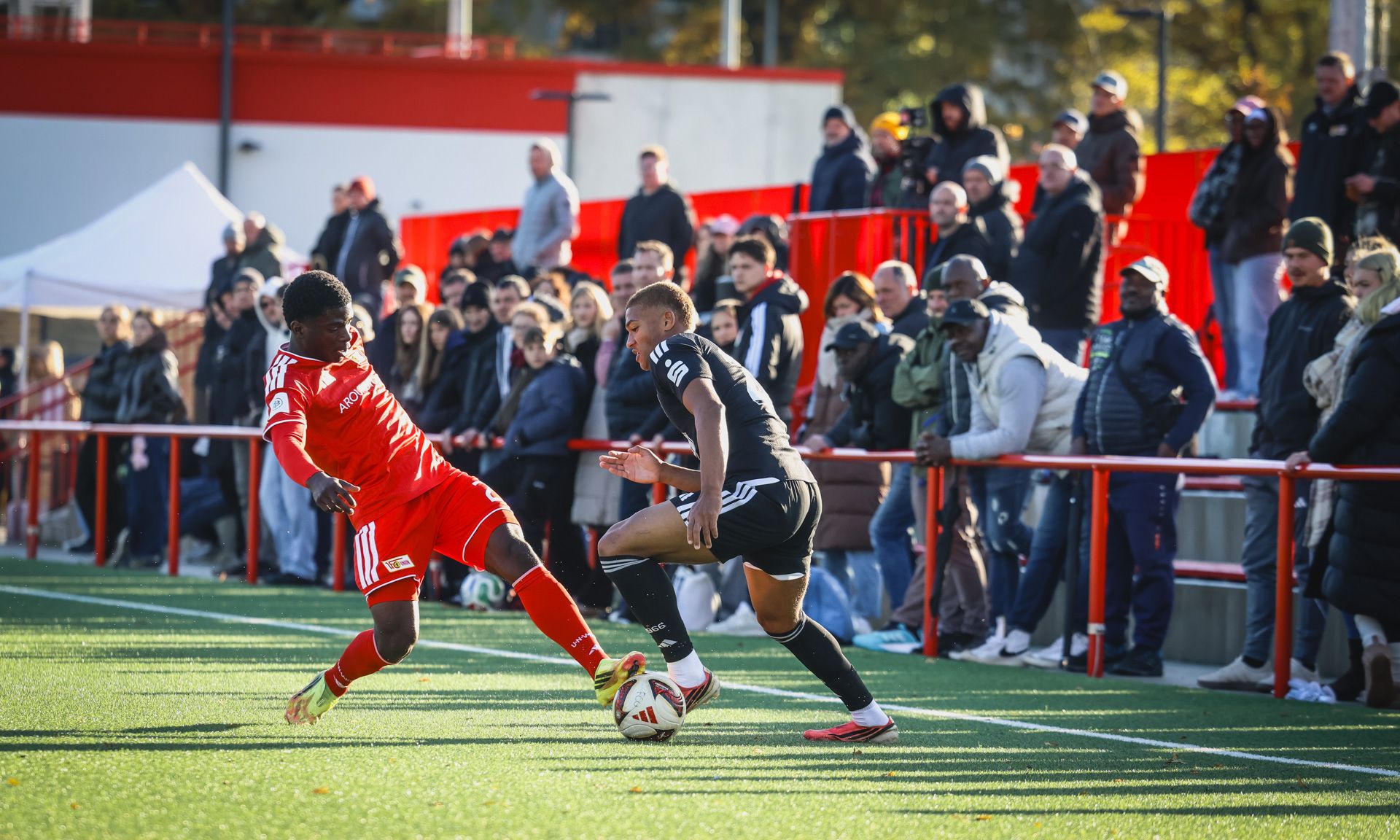Zwei Fußballspieler im Wettkampf um den Ball, mit Zuschauern im Hintergrund auf einer grünen Rasenfläche.