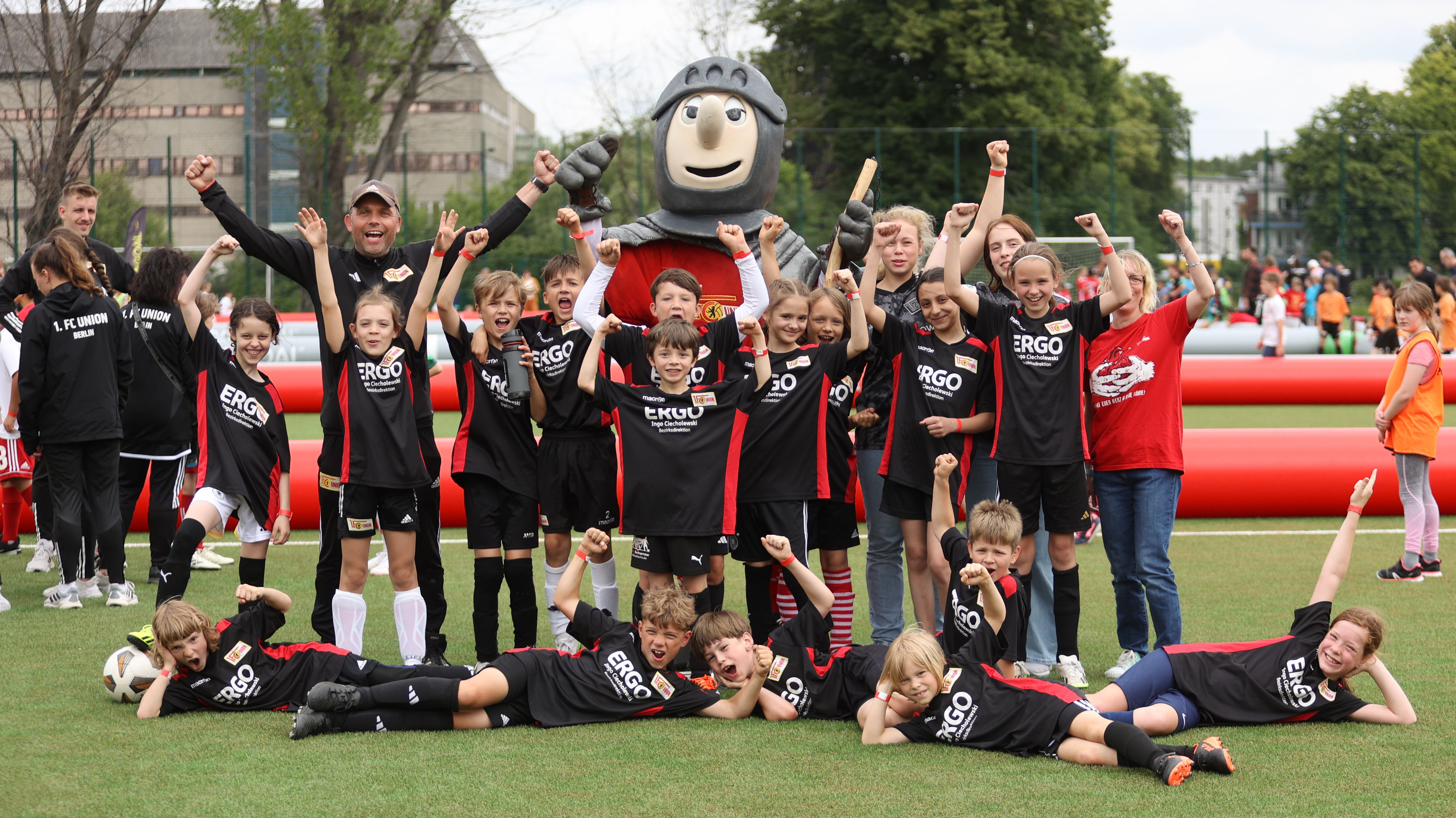Eine Gruppe von Kindern in Fußballtrikots posiert freudig mit einem Maskottchen auf einem Sportplatz.