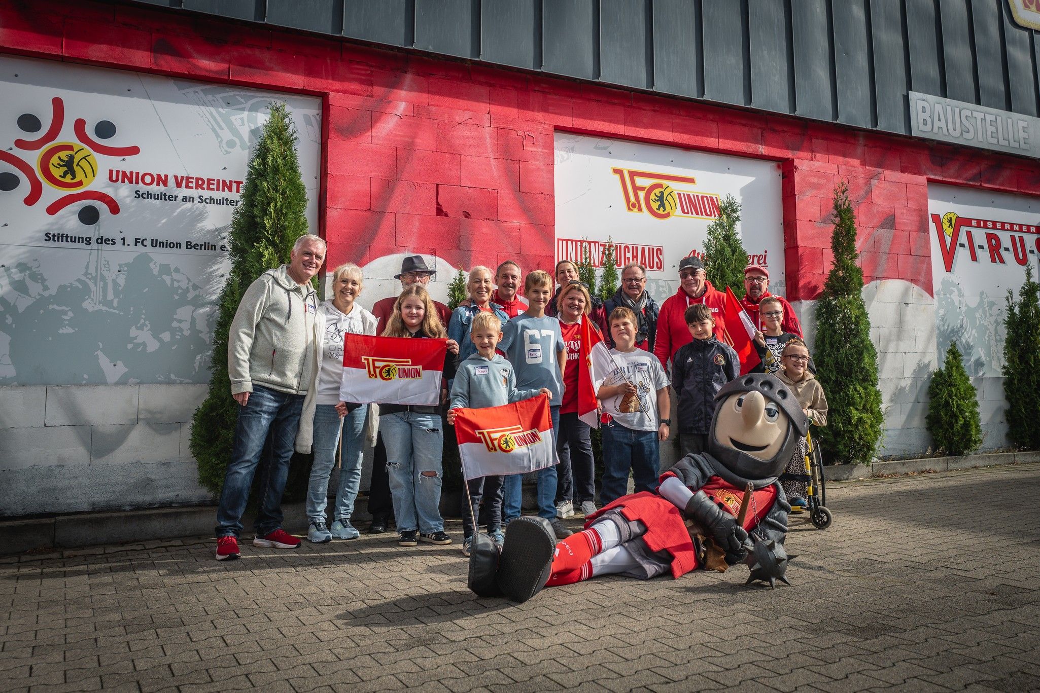 Gruppe von Menschen in sportlicher Kleidung, posierend mit einem Maskottchen vor einem roten Vereinsgebäude.
