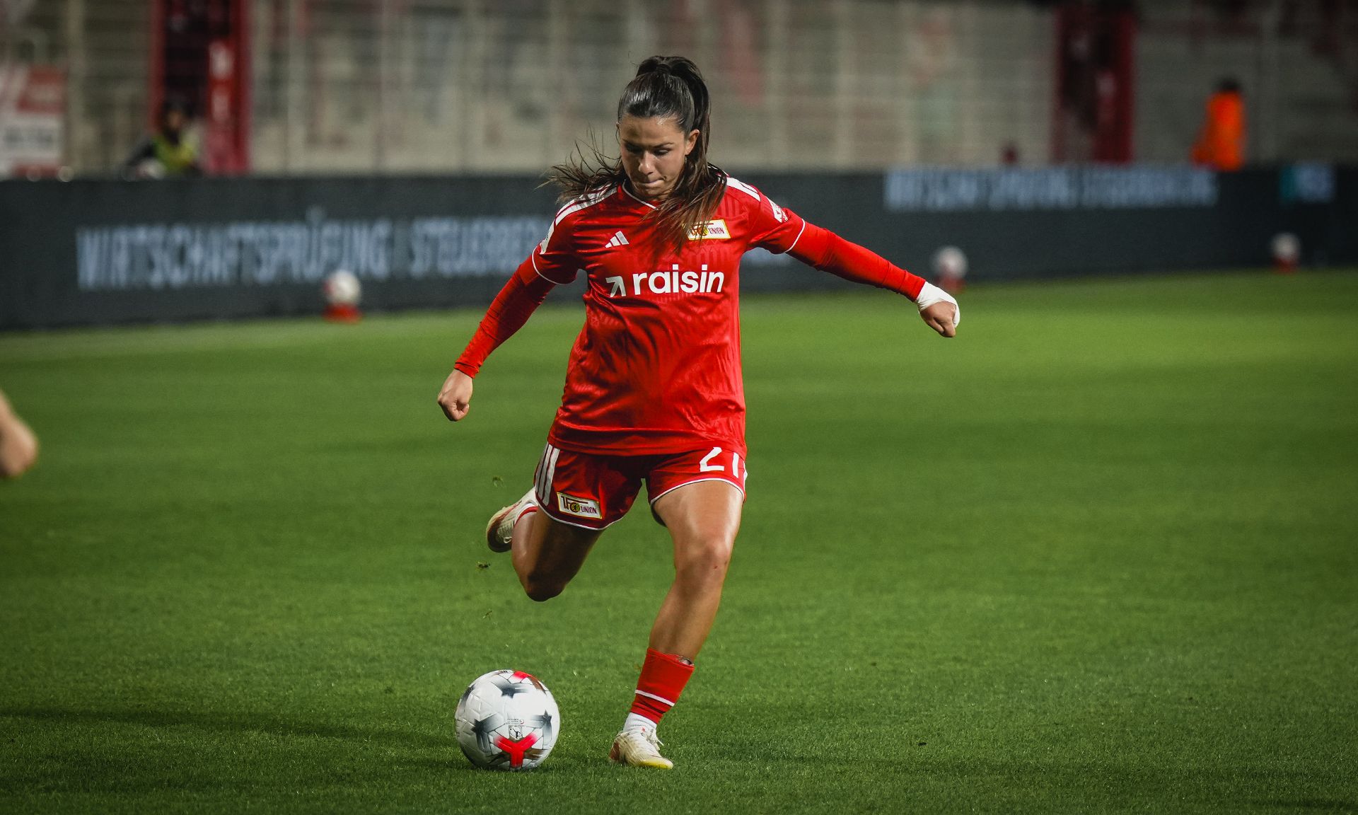 Eine Fußballspielerin in rotem Trikot tritt den Ball auf einem Rasenfeld.