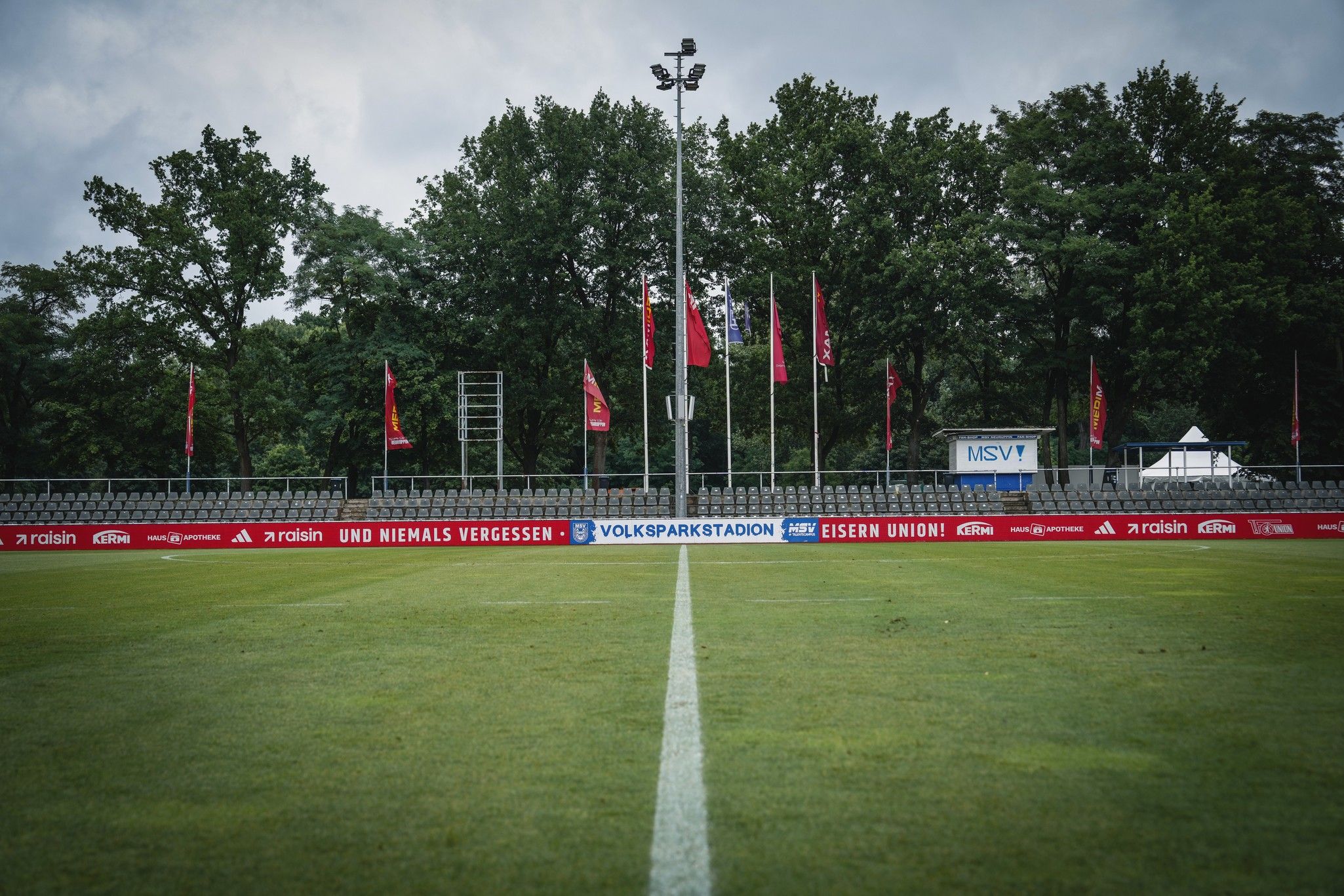 Eine grüne Fußballplatzmitte mit Linien, umgeben von Bänken und Fahnen unter bewölktem Himmel.