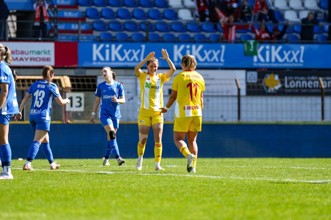 Spielerinnen eines Fußballteams feiern einen Treffer auf dem Feld, umgeben von Zuschauern und einer Stadionatmosphäre.