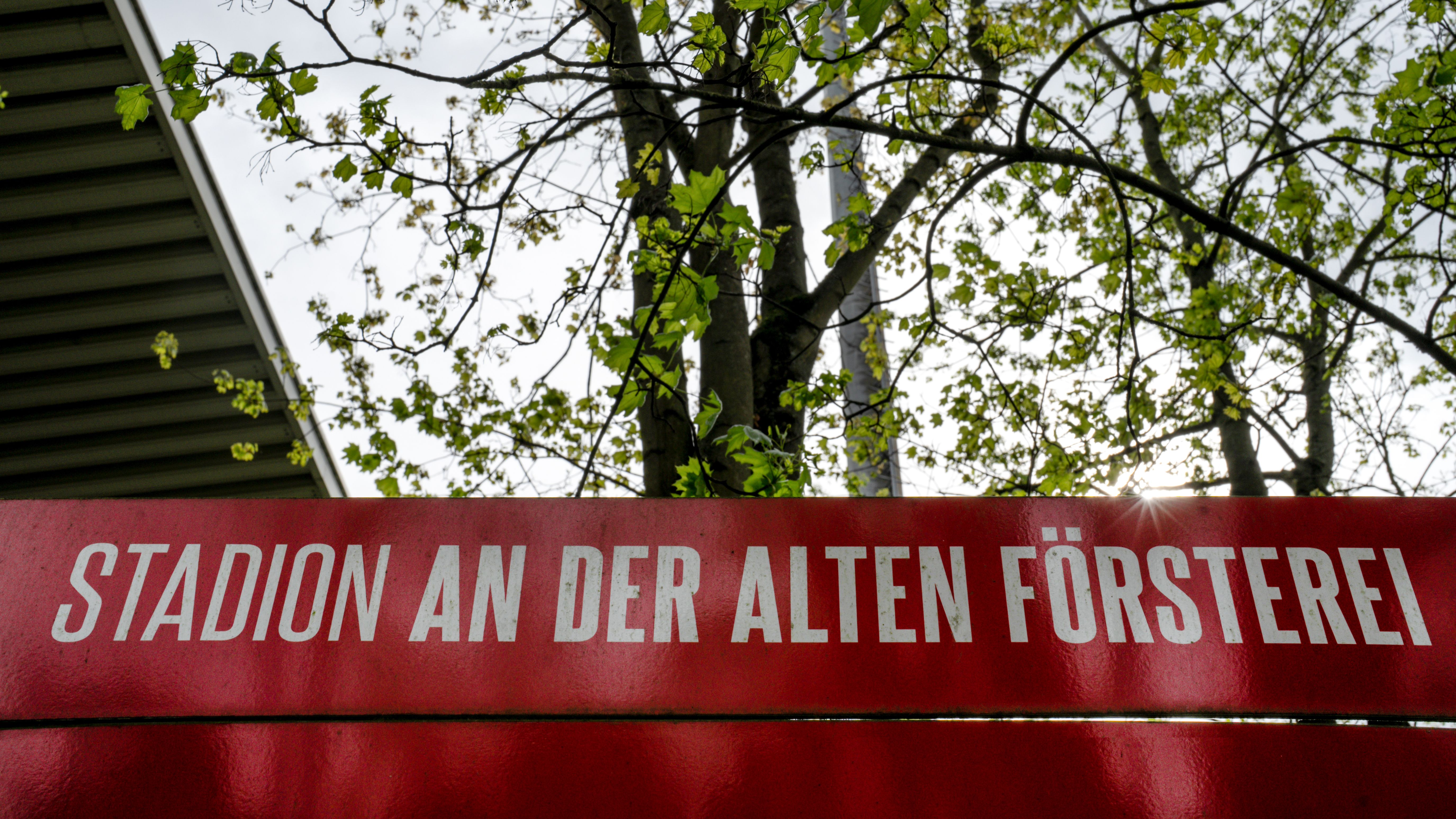 Schild mit der Aufschrift "Stadion an der alten Försterei" vor einem Baum und einer Gebäudefassade im Hintergrund.