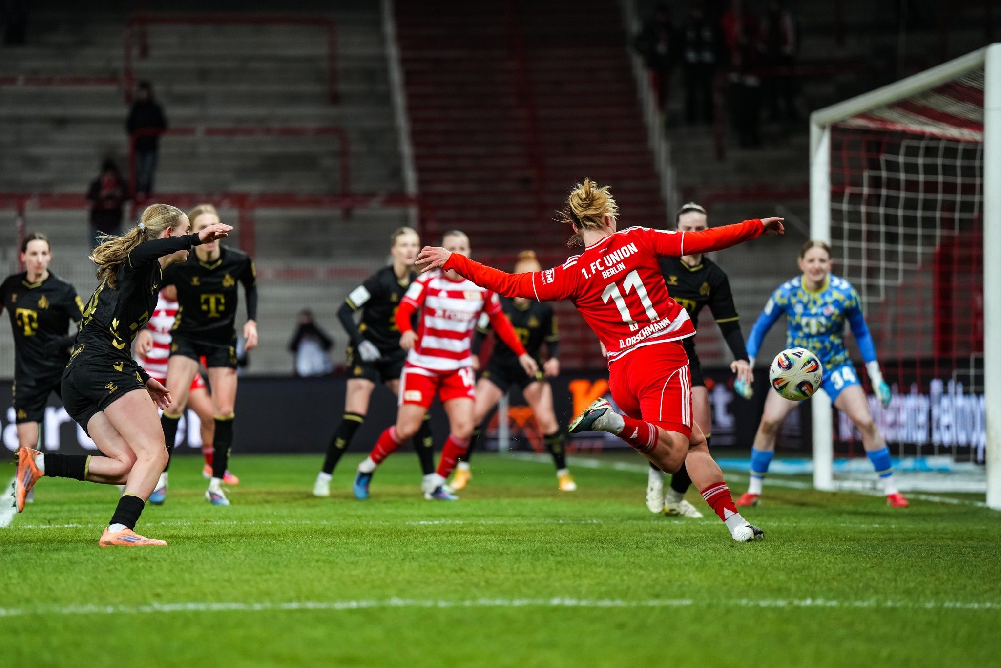 Eine Fußballspielerin in rotem Trikot schießt auf das Tor, während Mitspielerinnen in schwarzen Trikots und eine Torhüterin in blau zuschauen.