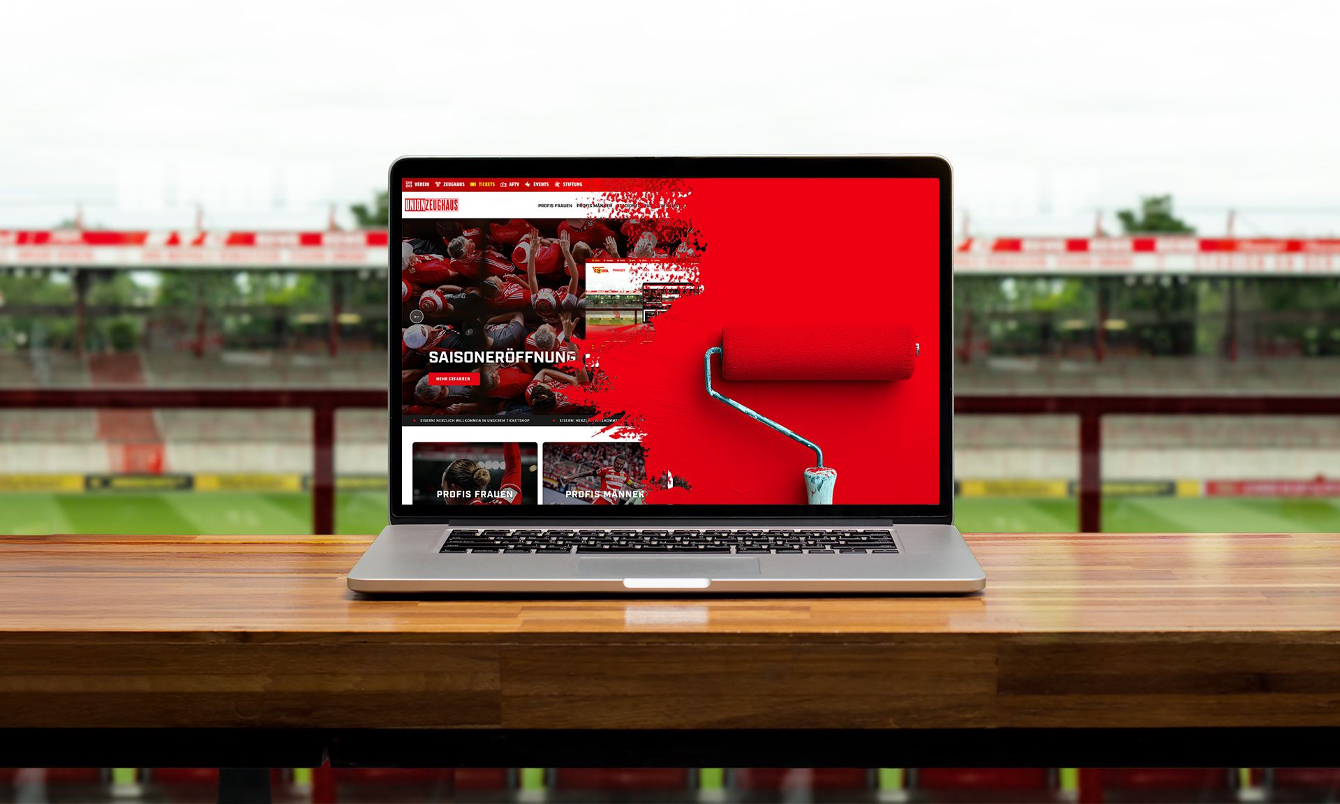 Laptop auf einem Tisch mit einem Fußballstadion im Hintergrund, zeigt eine rot gestaltete Webseite.