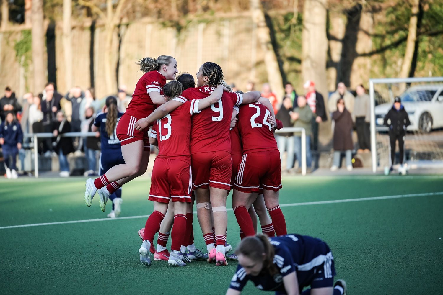 Eine Gruppe von Fußballspielerinnen in roten Trikots jubelt auf dem Feld, während eine Spielerin am Boden liegt.