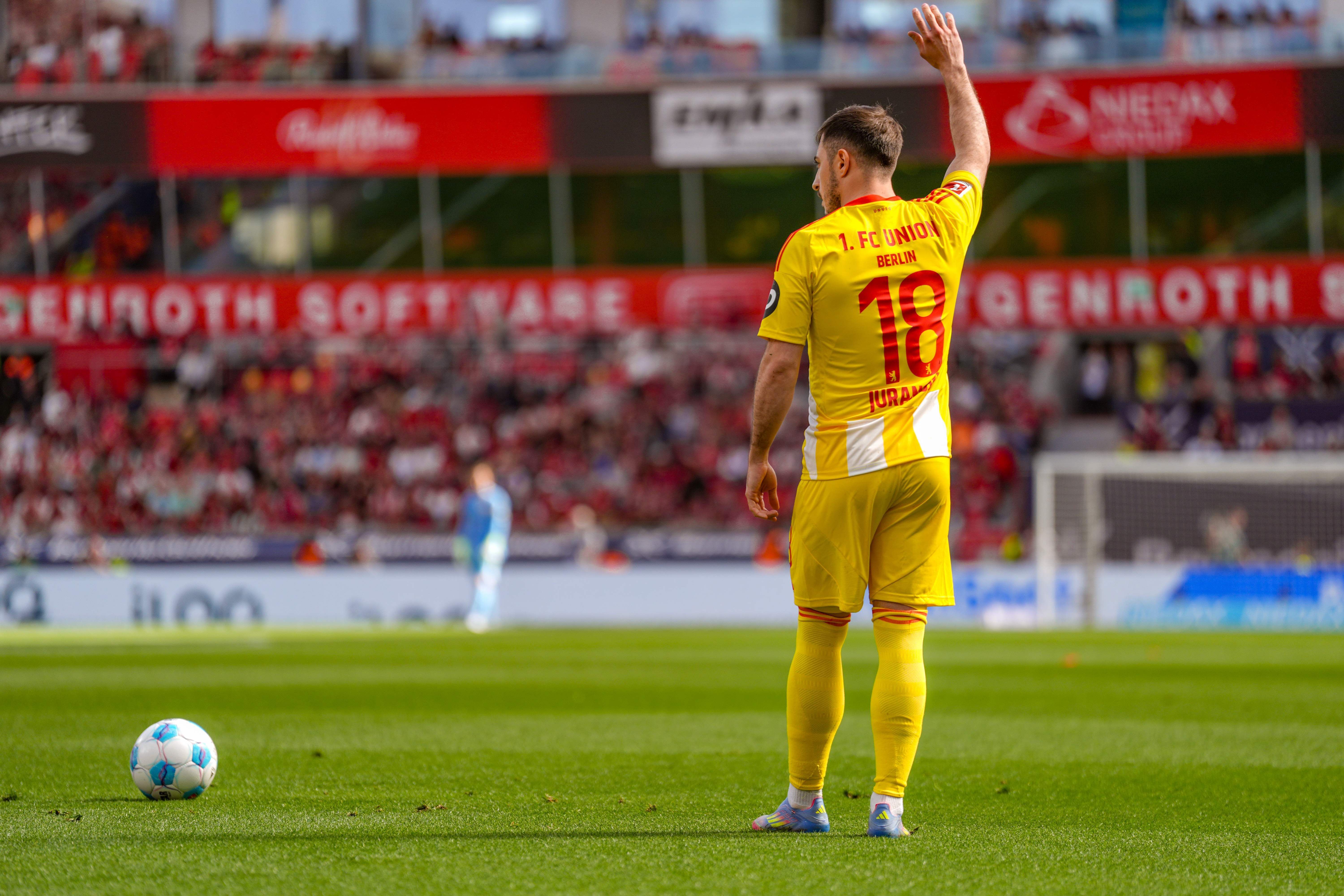 Ein Fußballspieler in gelbem Trikot mit der Nummer 18 hebt die Hand, während er auf dem Spielfeld steht. Fans sind im Hintergrund sichtbar.