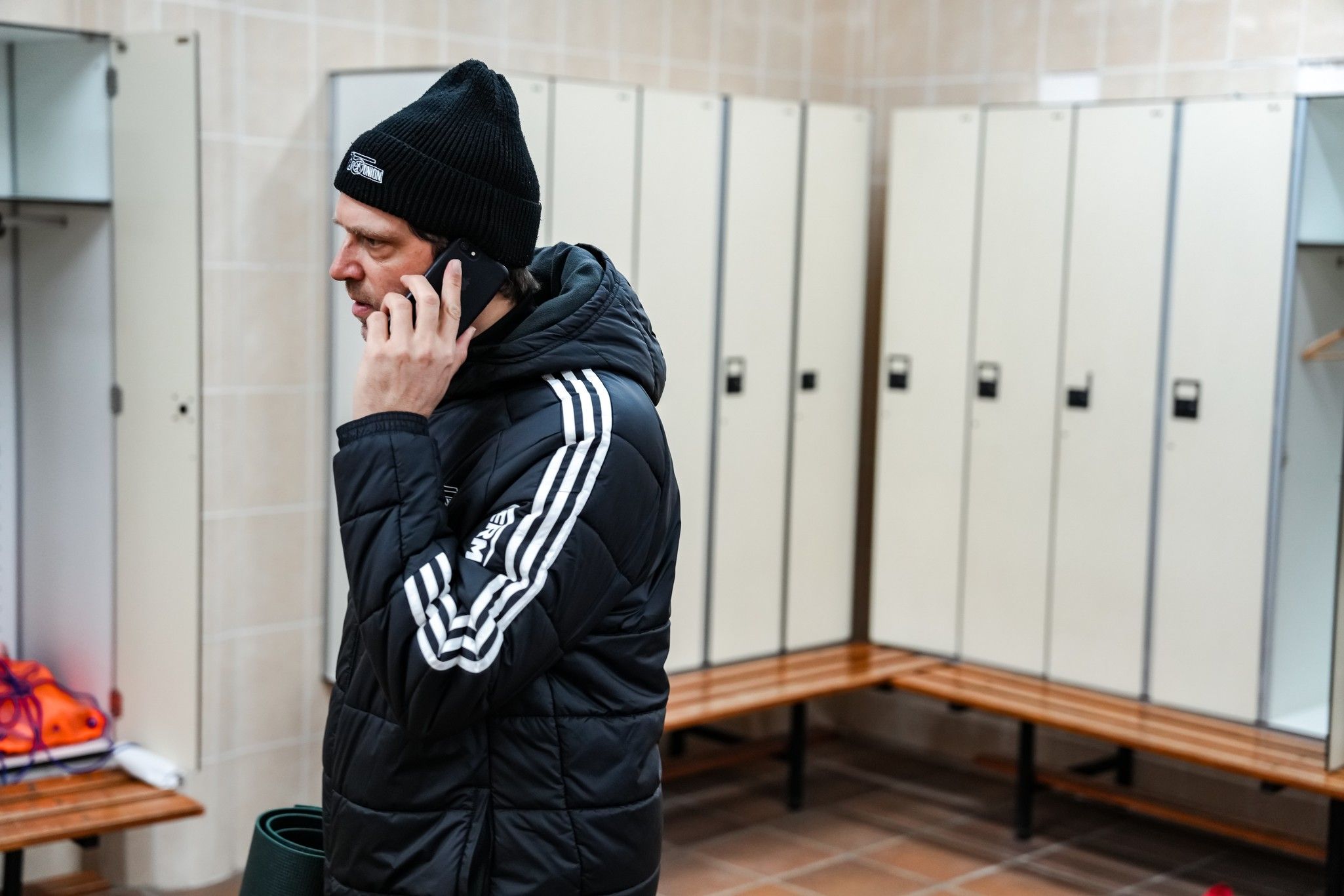 Ein Mann in einer Sportjacke mit Mütze spricht am Telefon in einem Umkleideraum mit Schließfächern.