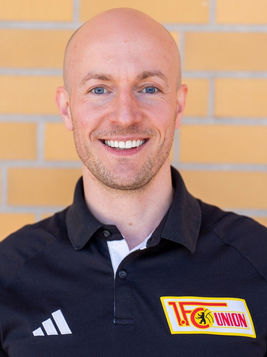 Eine lächelnde Person mit kurzer, glatzer Frisur trägt ein schwarzes Polohemd mit Logo von 1. FC Union. Hintergrund aus Ziegelsteinen.