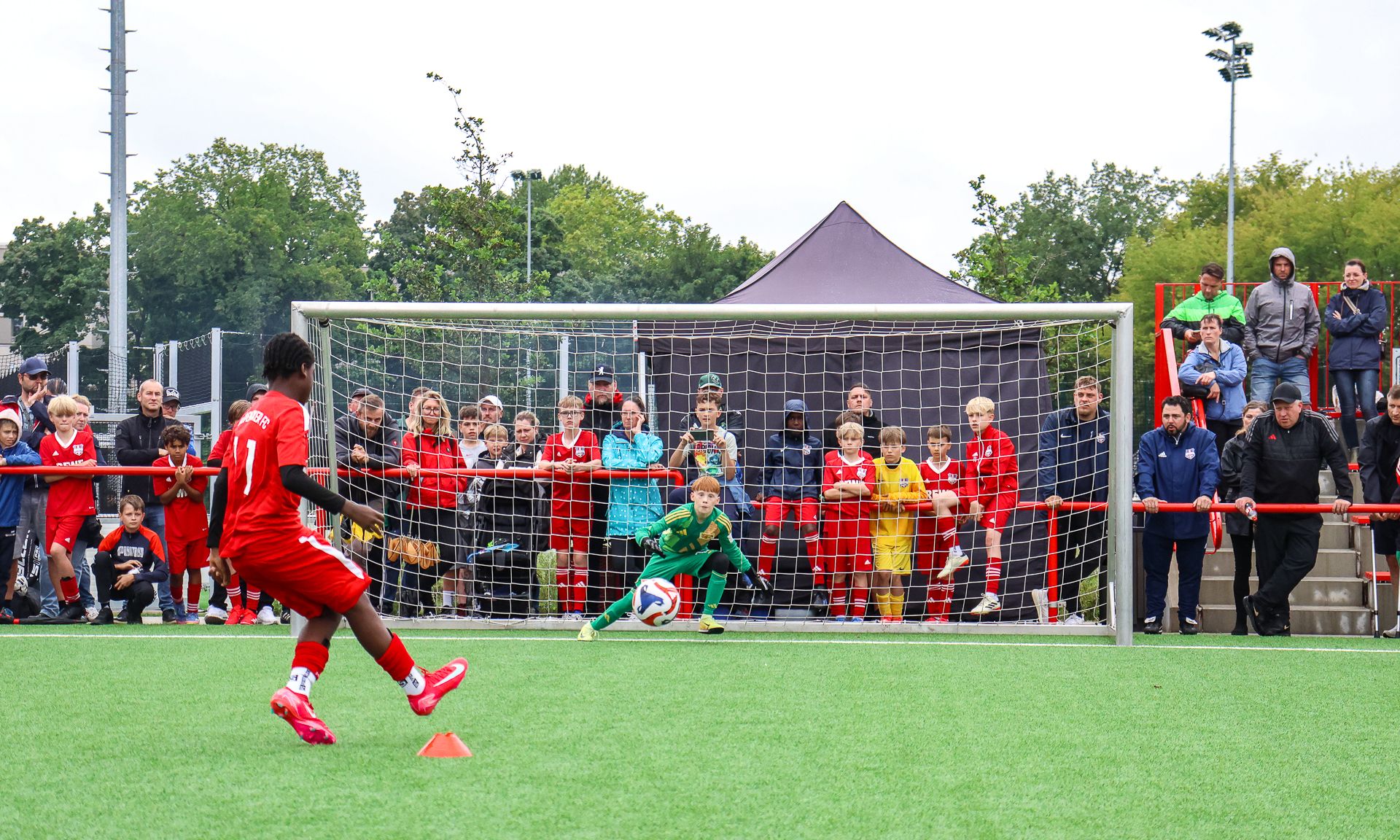 Fußballspiel mit einem Spieler beim Elfmeter und einem Torwart vor einer Zuschauermenge auf einem Kunstrasenplatz.