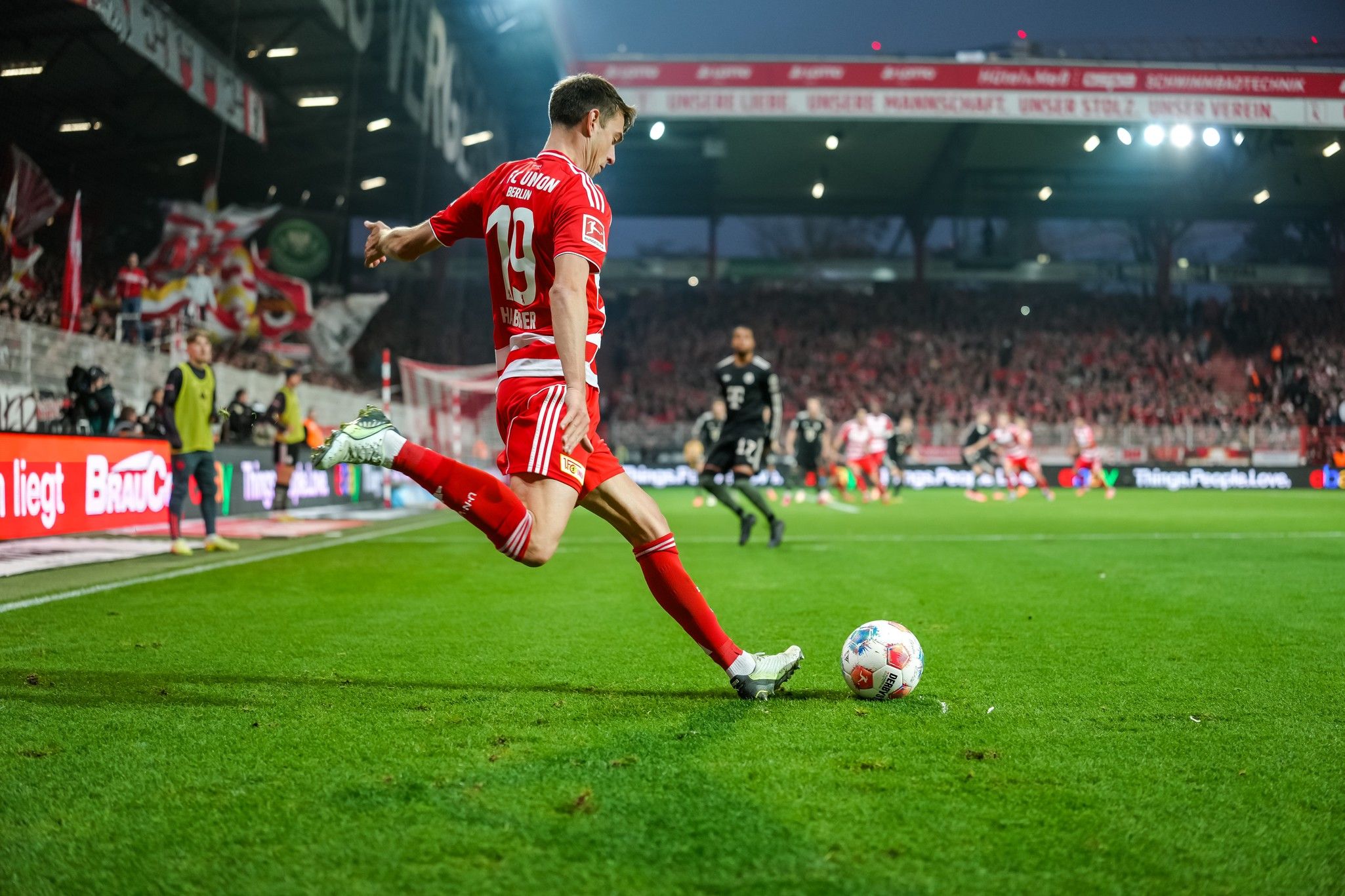 Ein Fußballspieler in rotem Trikot steht bereit, einen Eckball auf einem Stadionfeld mit jubelnden Fans im Hintergrund auszuführen.