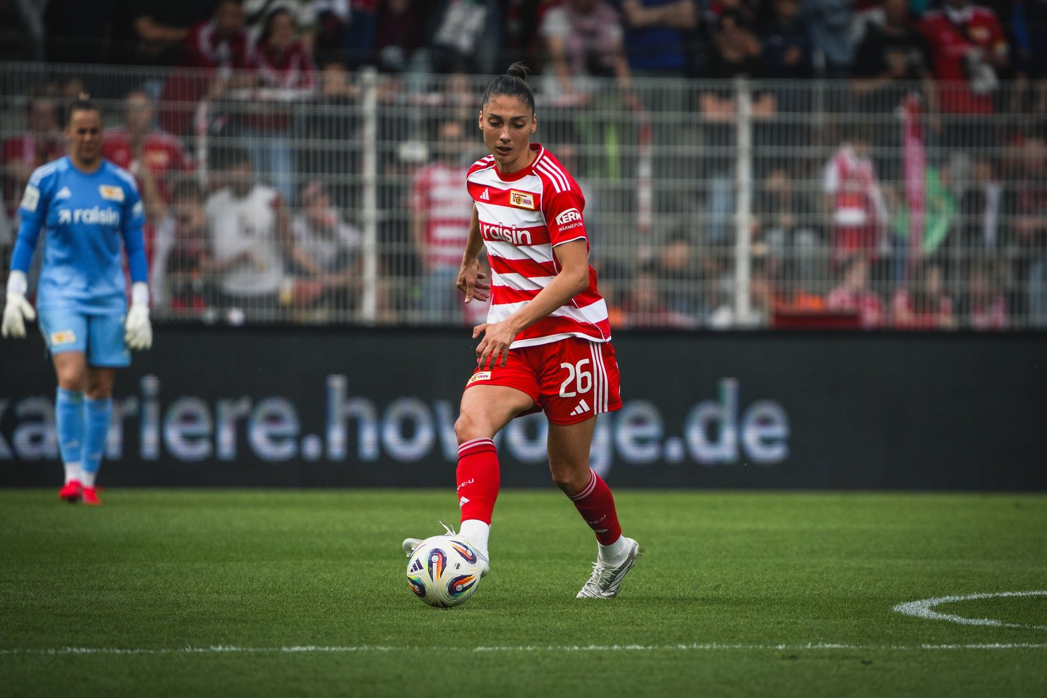 Spielerin in rotem Trikot mit der Nummer 26 dribbelt den Ball auf dem Fußballfeld, im Hintergrund ein Torwart.