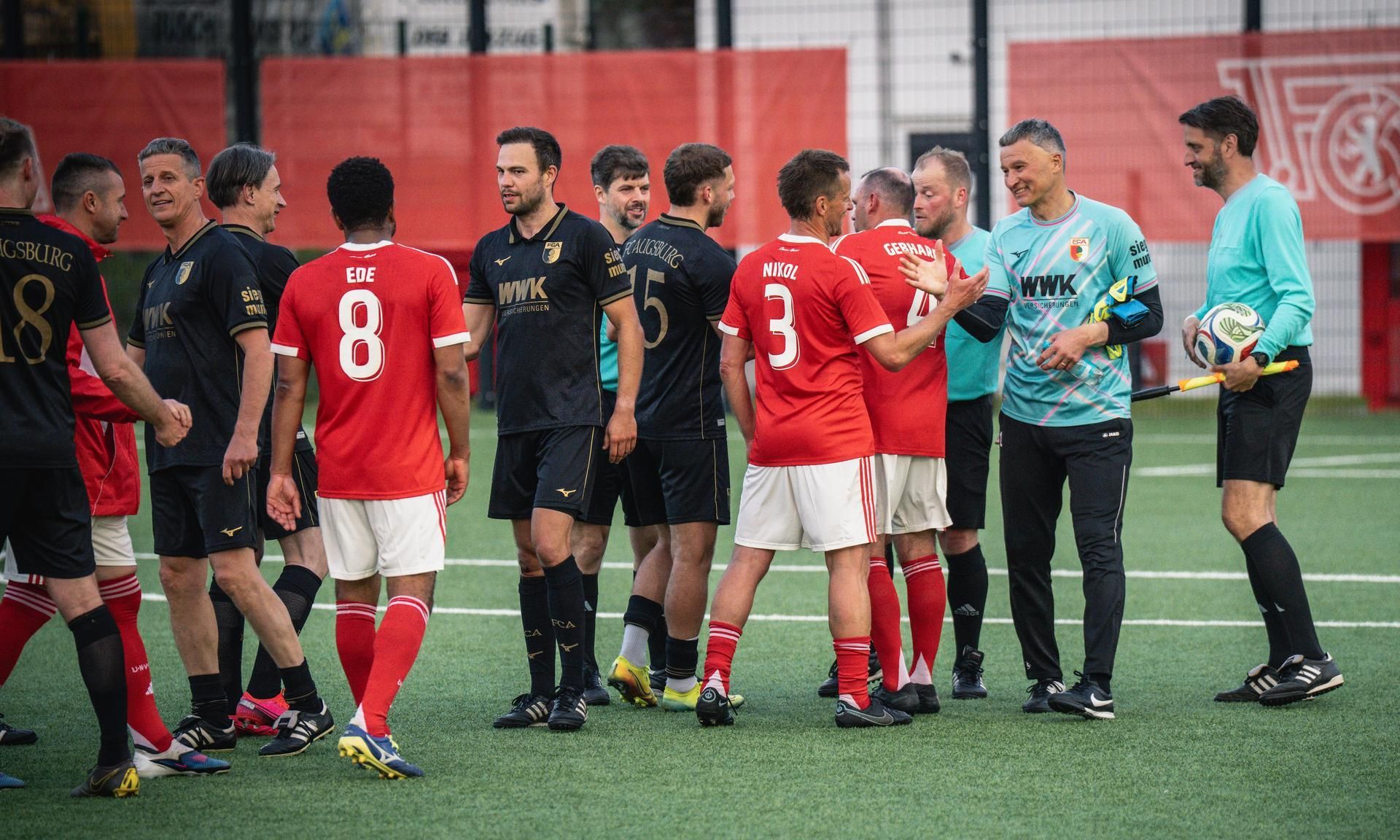 Gruppenszene von Fußballspielern in roten und schwarzen Trikots auf einem Platz, die sich nach dem Spiel begrüßen.