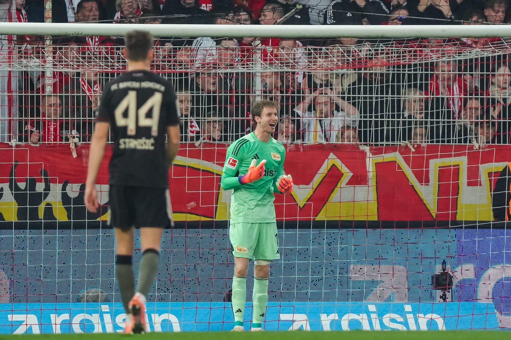 Ein Torwart in grüner Ausrüstung jubelt vor einem begeisterten Publikum, während ein Spieler der gegnerischen Mannschaft im Hintergrund steht.