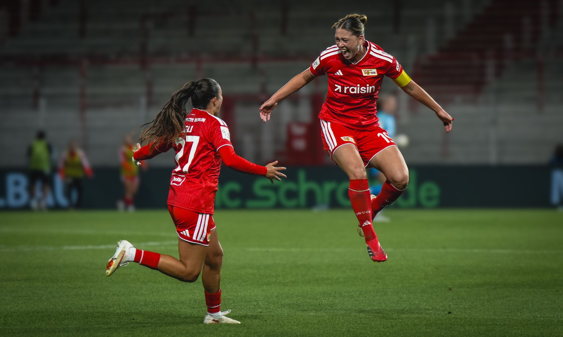 Zwei Fußballspielerinnen feiern ein Tor, eine springt jubelnd in die Luft, beide tragen rote Trikots des Vereins.