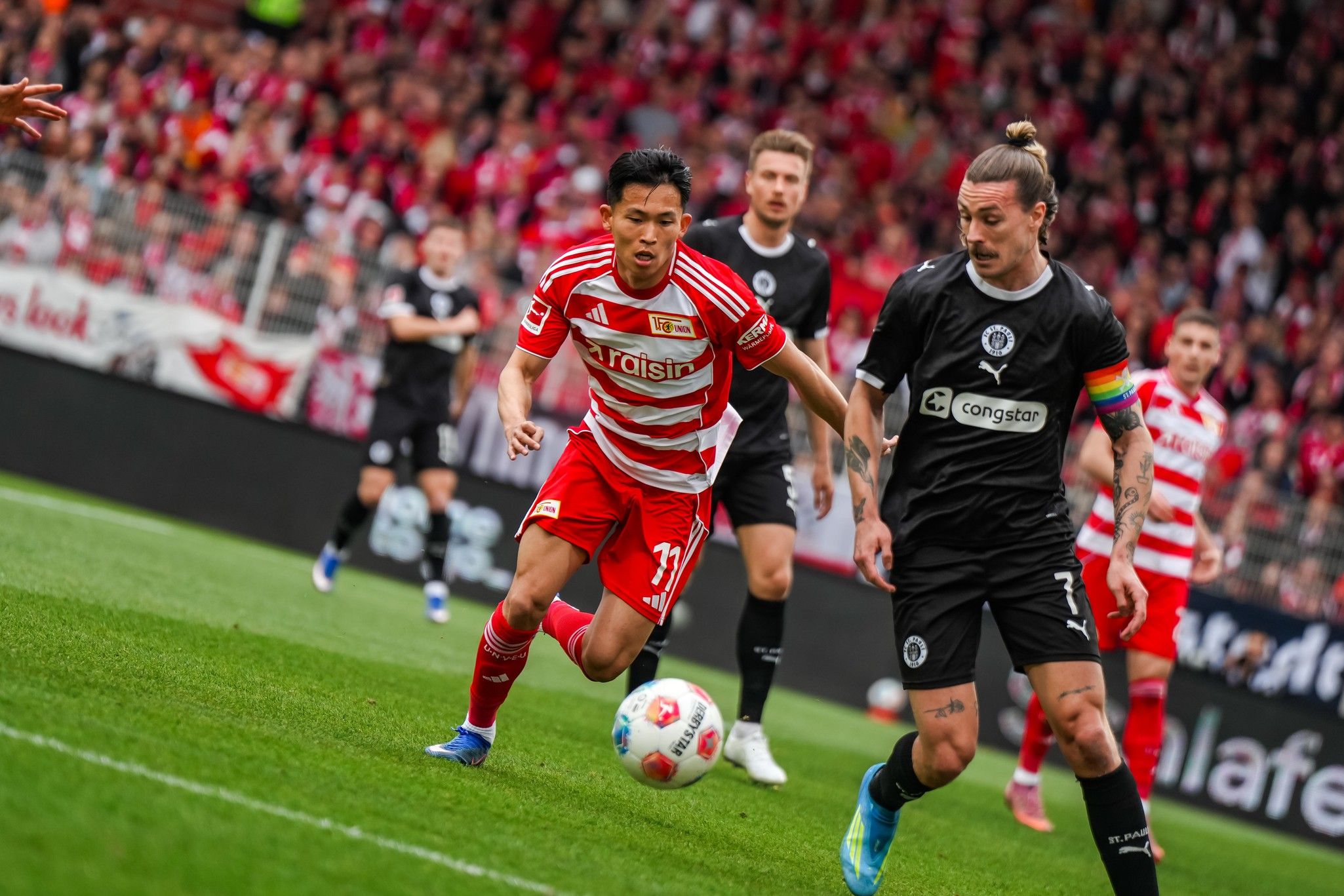 Ein Fußballspiel mit zwei Spielern in Aktion, einer in rot-weißer Uniform und der andere in schwarz, umgeben von Fans im Hintergrund.