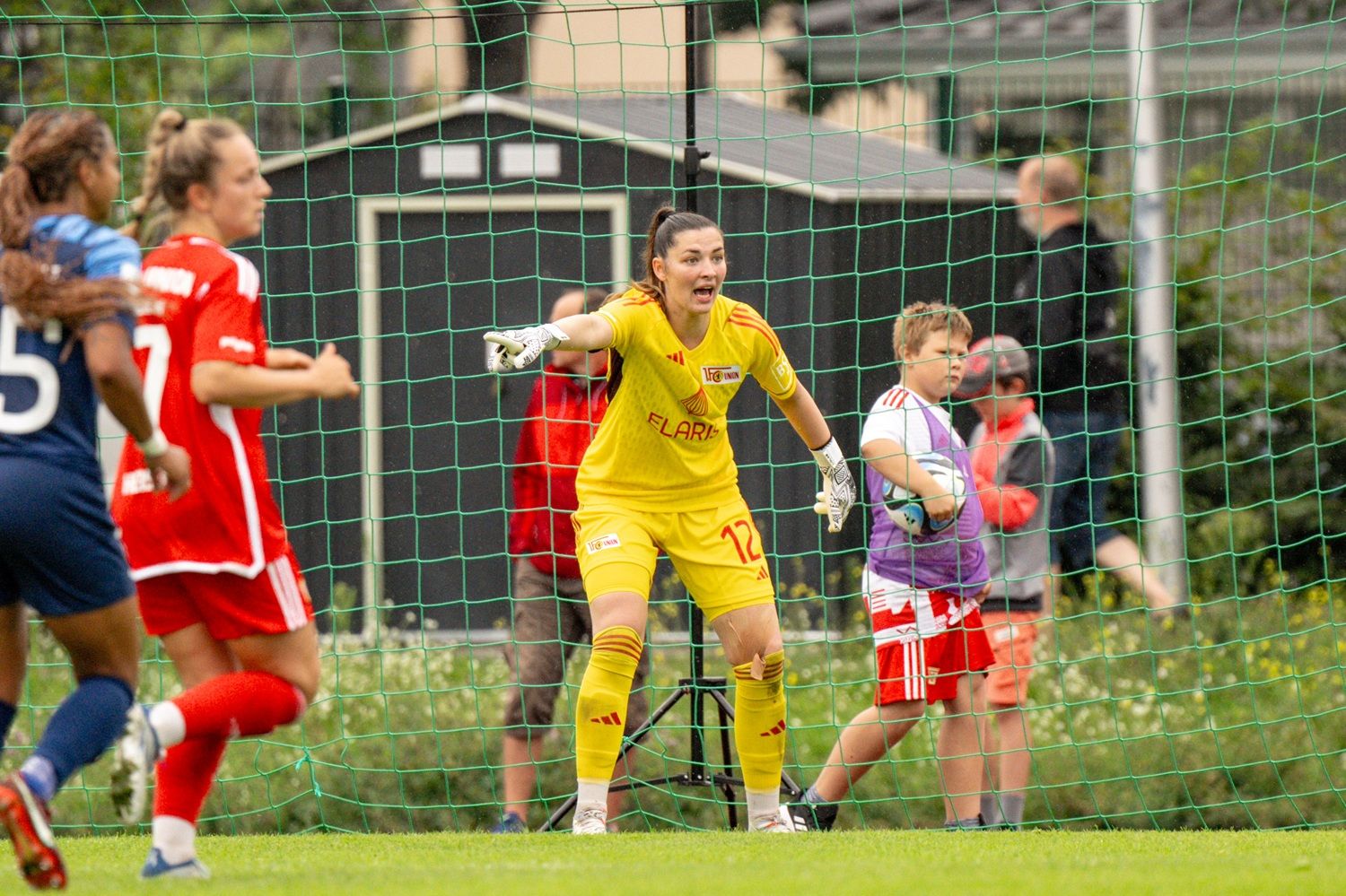 Torwartin in gelber Ausrüstung spricht auf dem Spielfeld, während Spielerinnen in roten und blauen Trikots im Hintergrund agieren.