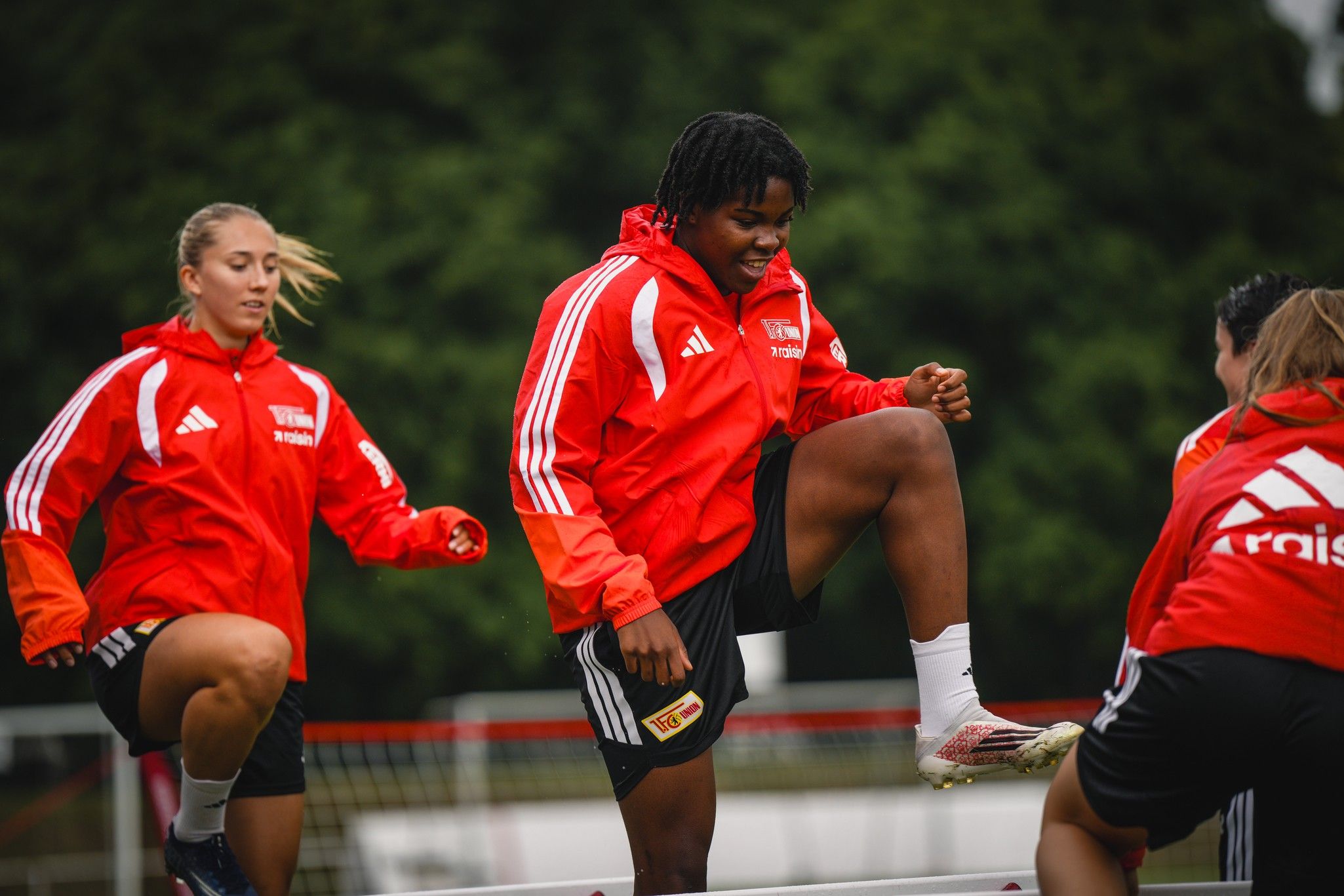 Drei Fußballspielerinnen in roten Trainingsjacken üben in einem Stadion, während sie auf eine Trainingsstation steigen.