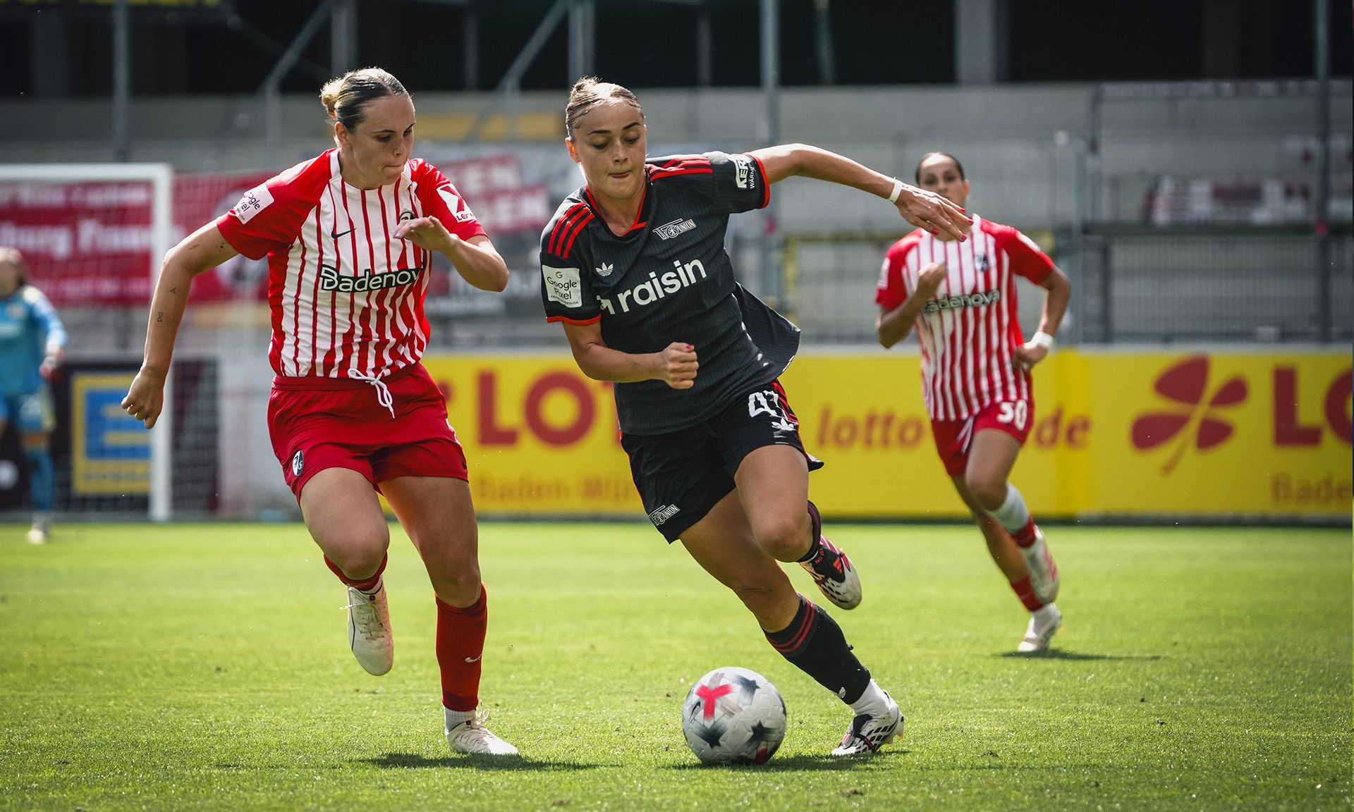 Zwei Fußballspielerinnen im sprintenden Duell auf dem Feld, eine trägt ein schwarzes Trikot mit der Nummer 47, die andere ein gestreiftes Trikot.