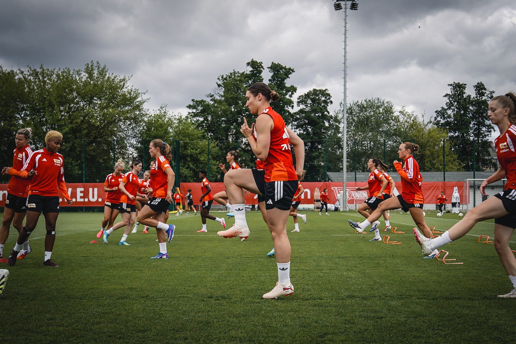 Frauenfußballmannschaft beim Training, sie führen Aufwärmübungen auf einem Rasenplatz unter bewölktem Himmel durch.