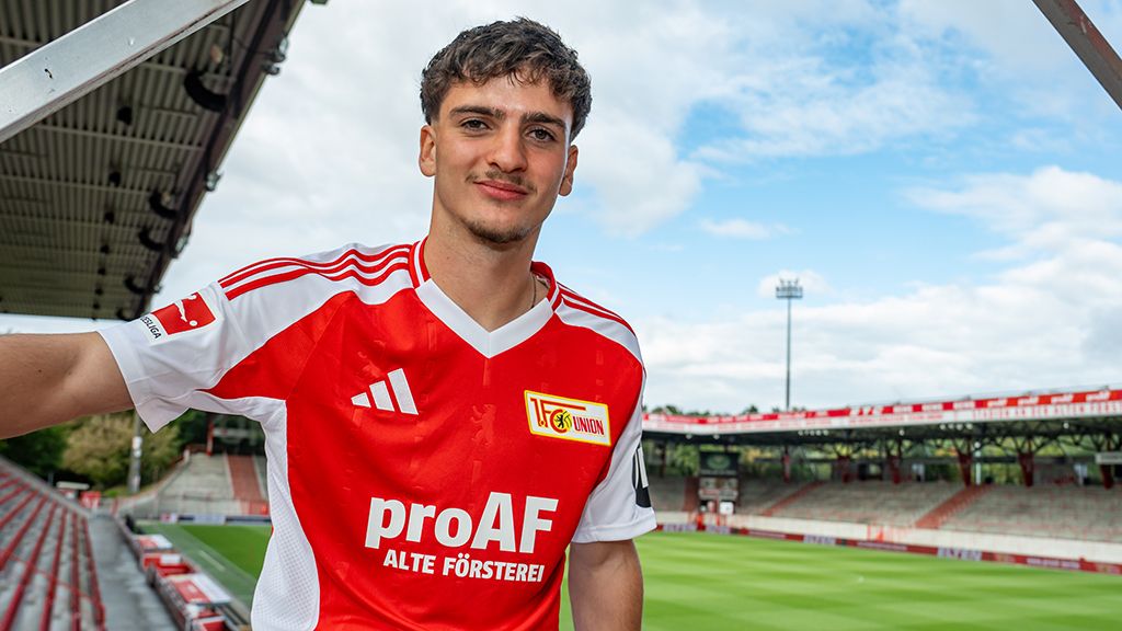 Junger Fußballspieler im rot-weißen Trikot steht lächelnd im Stadion, im Hintergrund das Spielfeld und der Himmel.