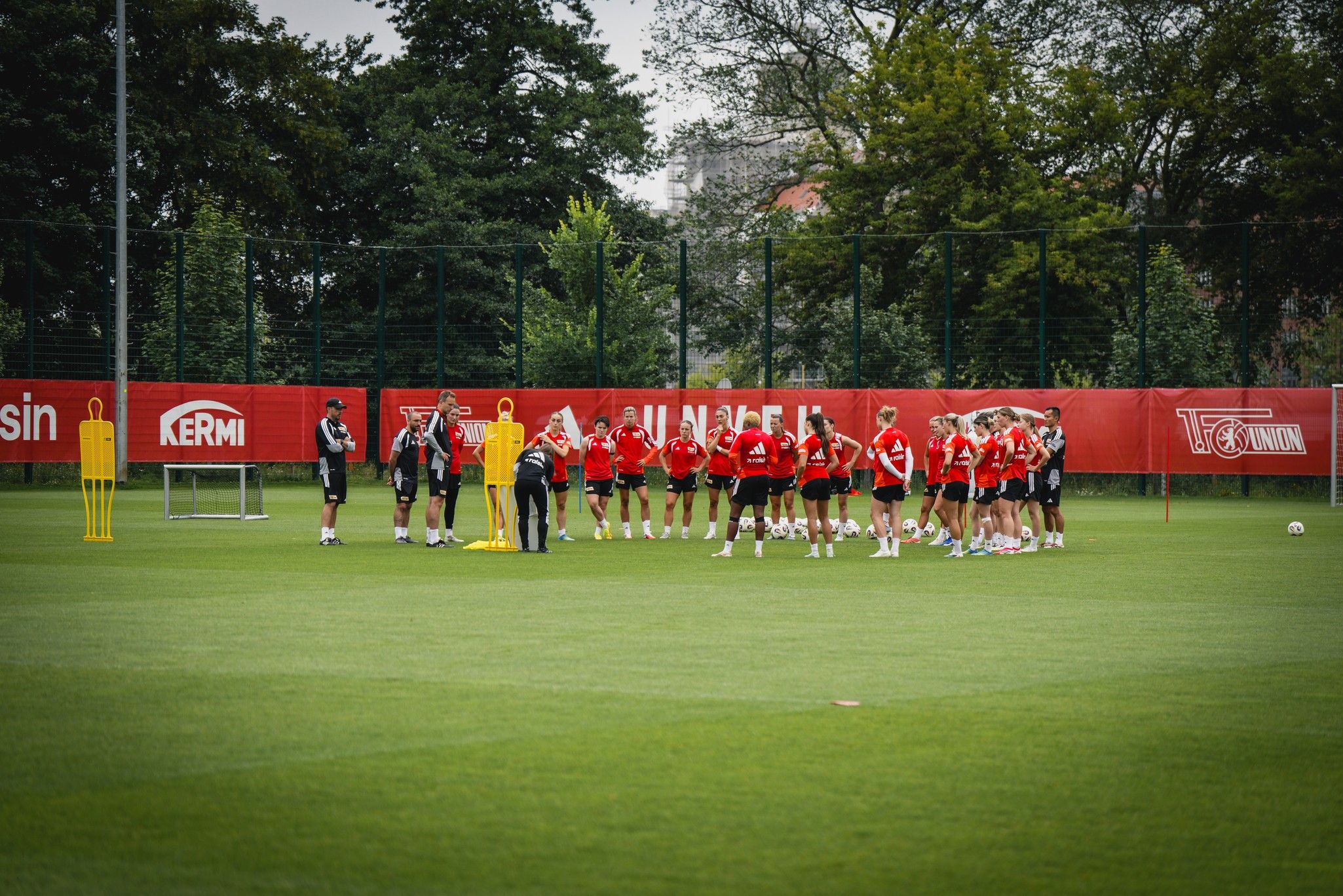 Eine Gruppe von Fußballspielern in roten Trikots versammelt sich auf dem Trainingsfeld um einen Trainer. Im Hintergrund sind Bäume und ein Zaun.