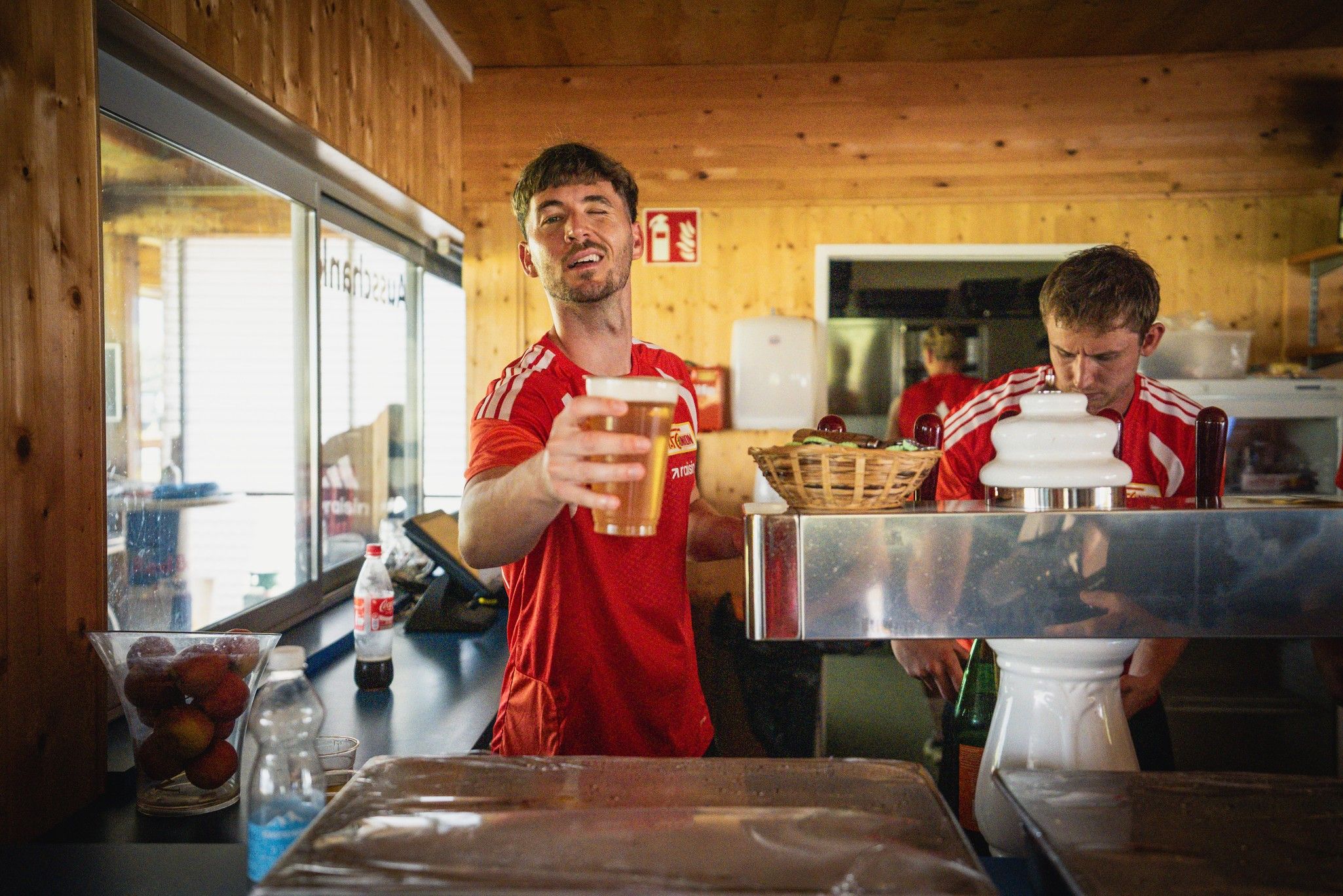 Zwei Personen in roten Shirts stehen hinter einer Bar, einer hält ein Bierglas, während der andere beschäftigt ist.