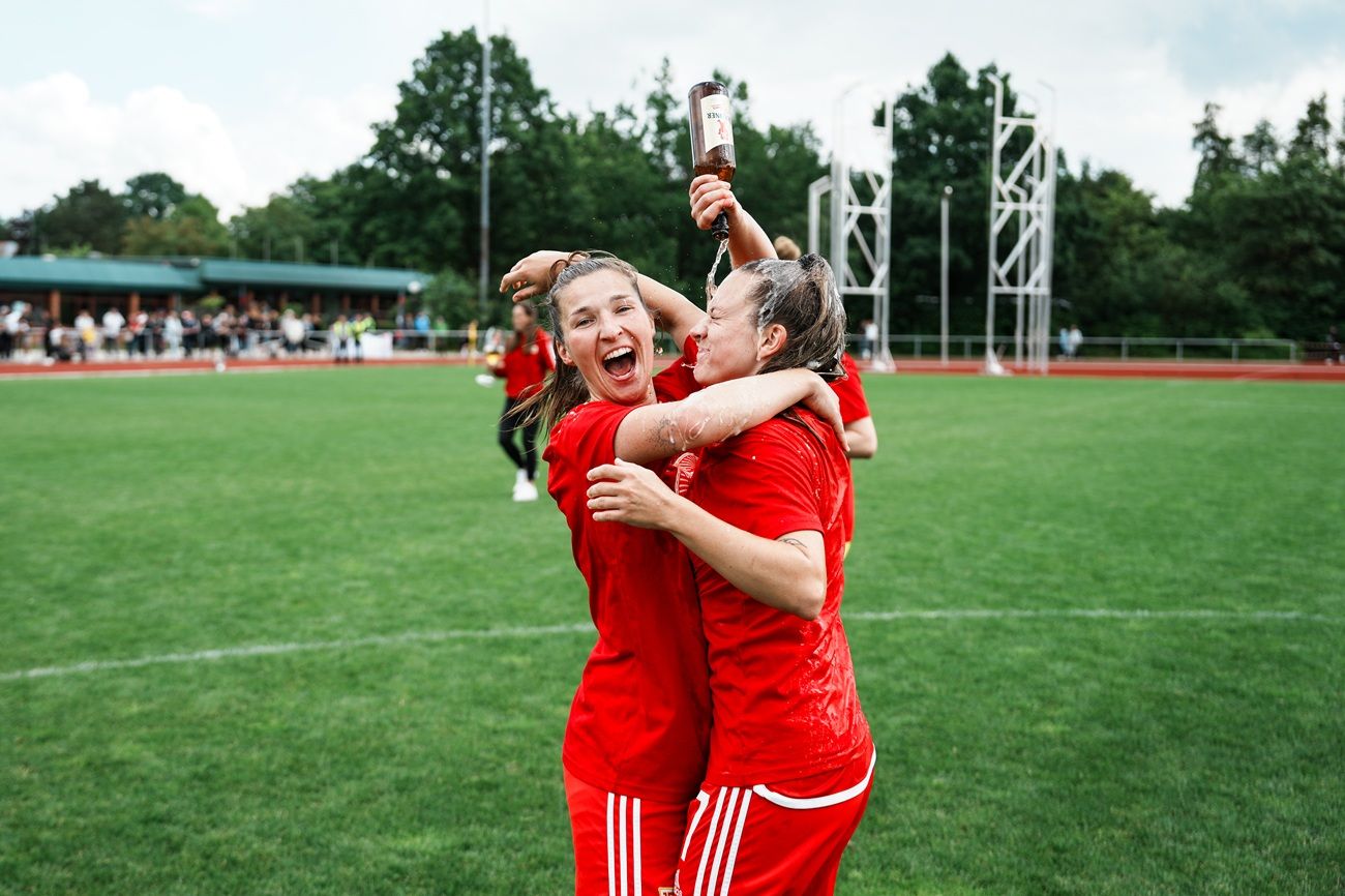 Zwei Spielerinnen in roten Trikots umarmen sich freudig auf einem Fußballfeld, umgeben von jubelnden Fans.