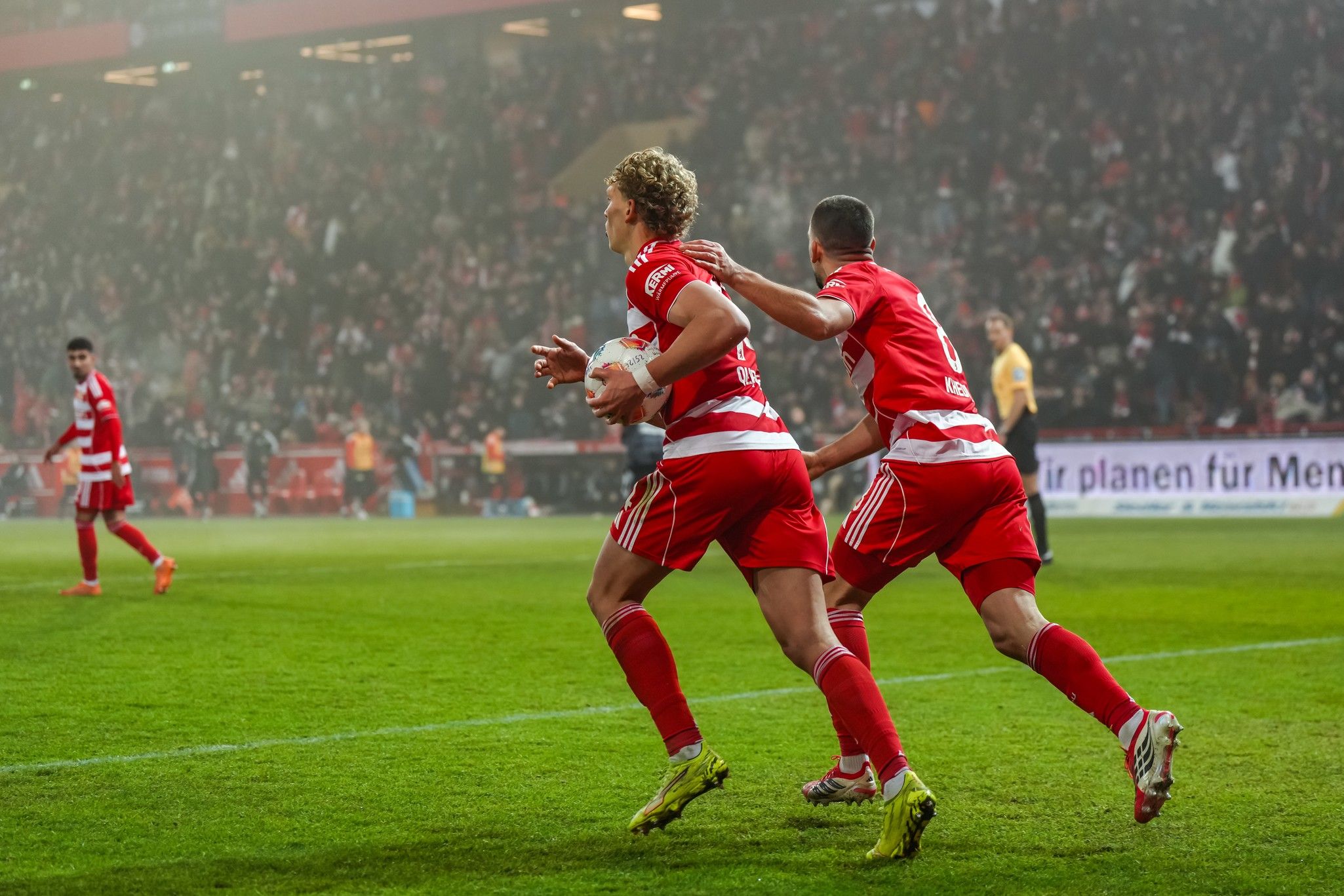 Spieler in roten Trikots feiern einen Torerfolg auf einem Fußballfeld vor jubelnden Fans im Hintergrund.
