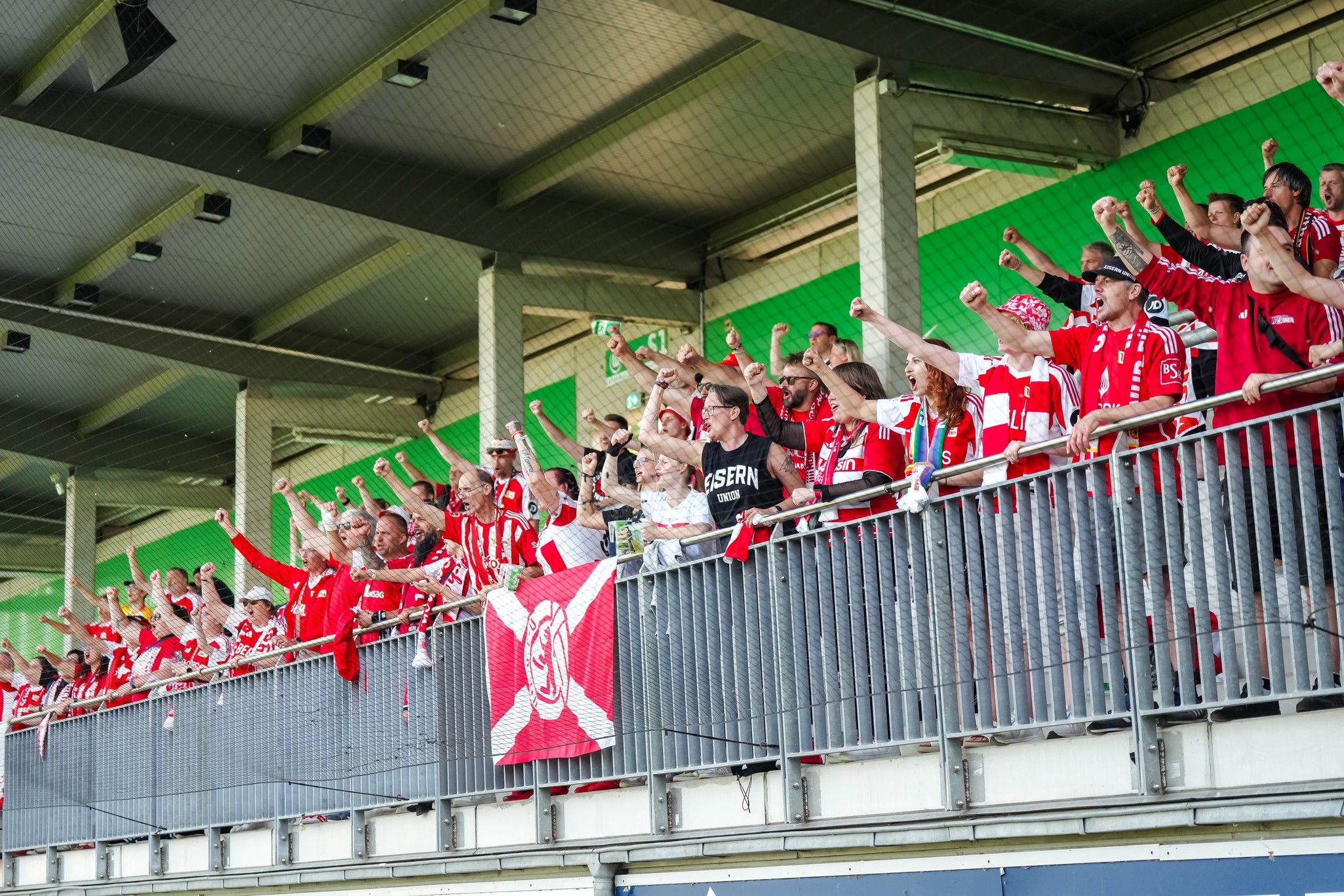Eine große Gruppe von Fans in Rot und Weiß jubelt und hebt die Hände, während sie in einem Stadion stehen.