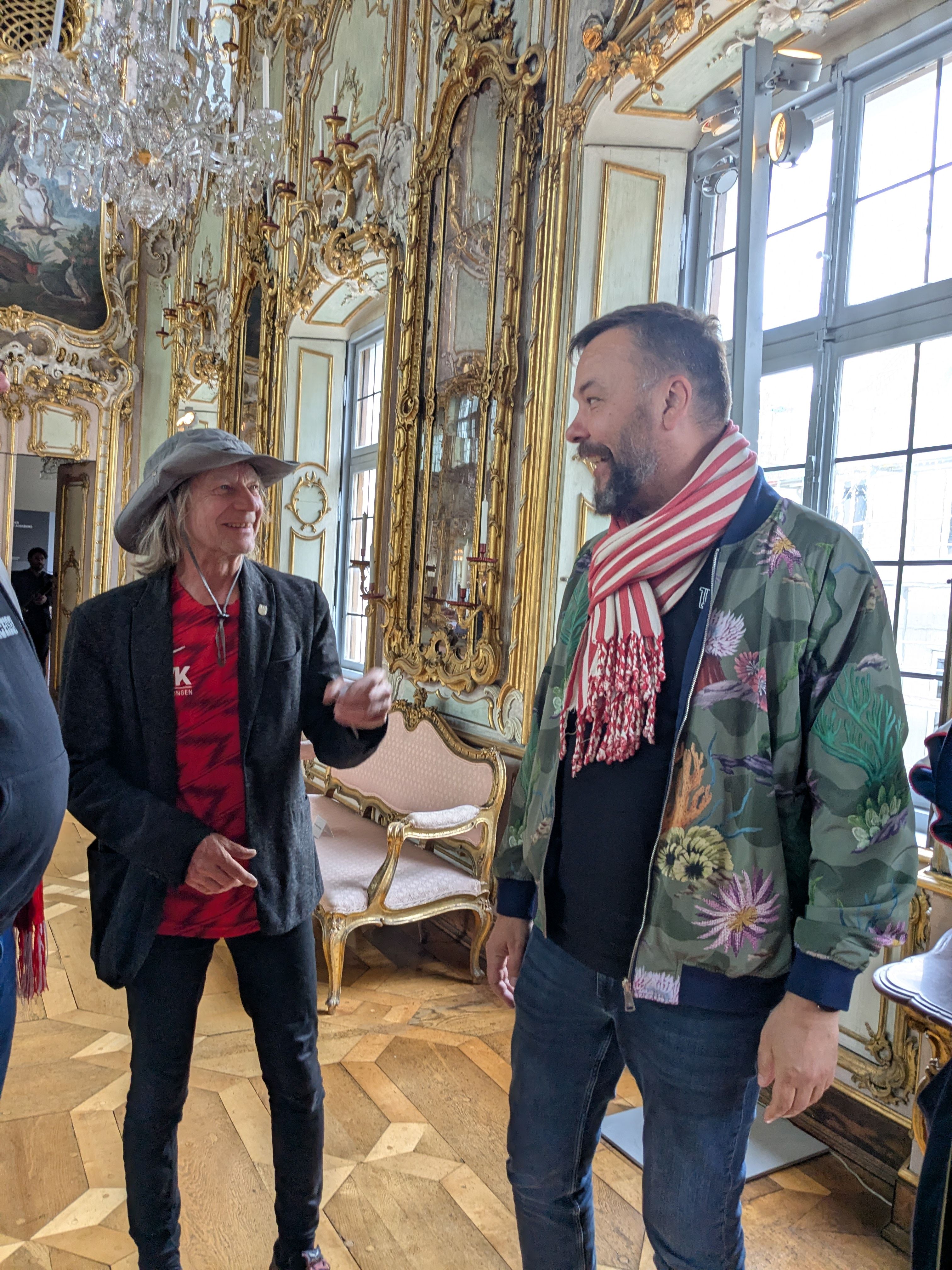 Zwei Männer unterhalten sich in einem prächtigen Raum mit Barockdekor und großen Fenstern.