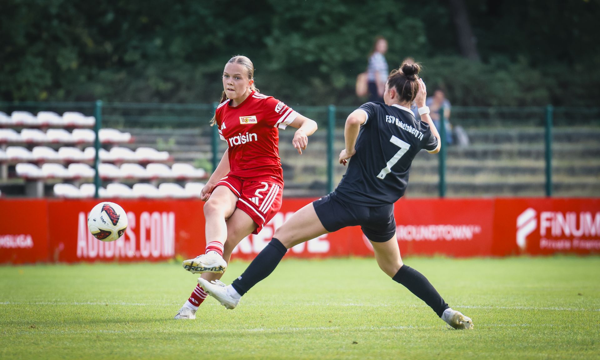 Eine Fußballspielerin in rotem Trikot schießt den Ball, während eine gegnerische Spielerin in Schwarz versucht, den Ball zu blocken.