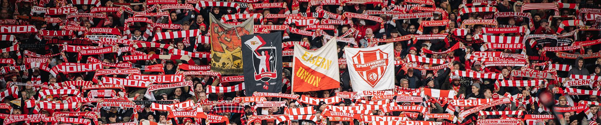 Eine große Menschenmenge im Stadion hält rote und weiße Schals hoch und zeigt Fahnen mit Vereinslogos.