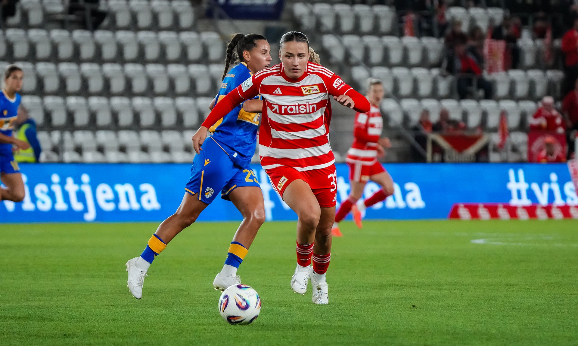 Eine Spielerin im rot-gestreiften Trikot dribbelt den Ball, während eine gegnerische Spielerin in gelb-blauem Trikot追いかける.