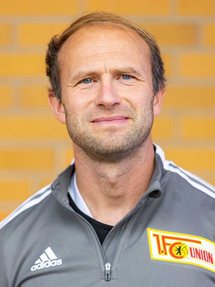 Ein lächelnder Mann mit kurzen braunen Haaren, trägt ein graues Trainingsoutfit mit dem Logo eines Sportvereins im Hintergrund.