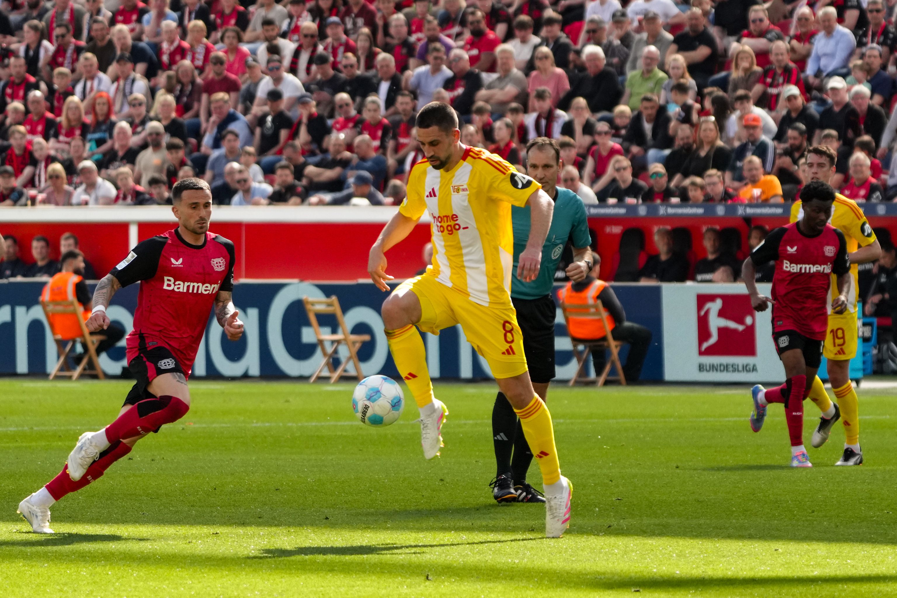 Ein Fußballspieler in gelber und roter Trikotausführung dribbelt den Ball, während ein Gegner in rotem Trikot ihn verfolgt.
