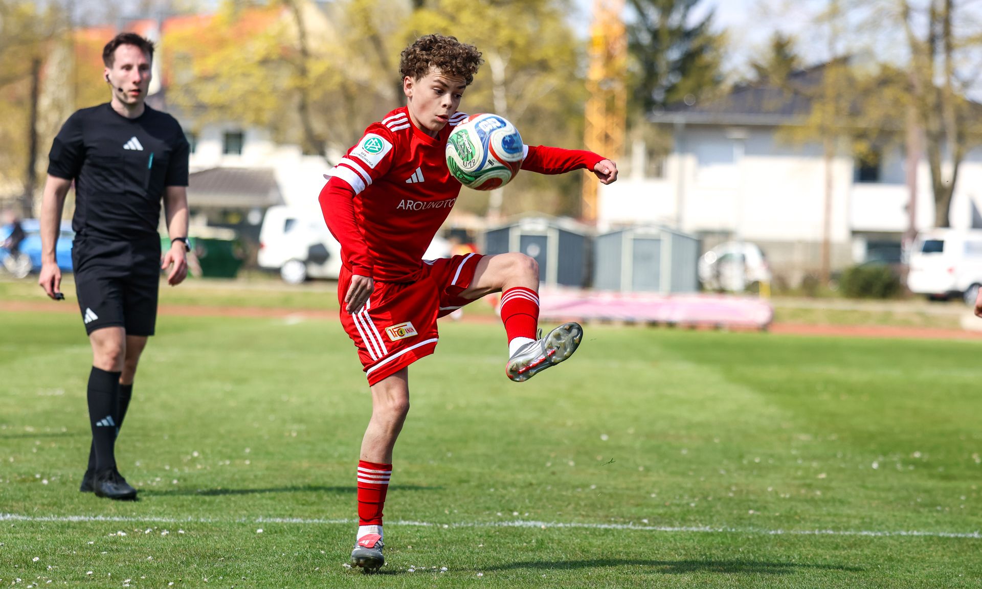 Ein junger Fußballspieler in roter Kleidung dribbelt einen Ball auf dem Feld, während ein Gegner in schwarzer Kleidung zuschaut.