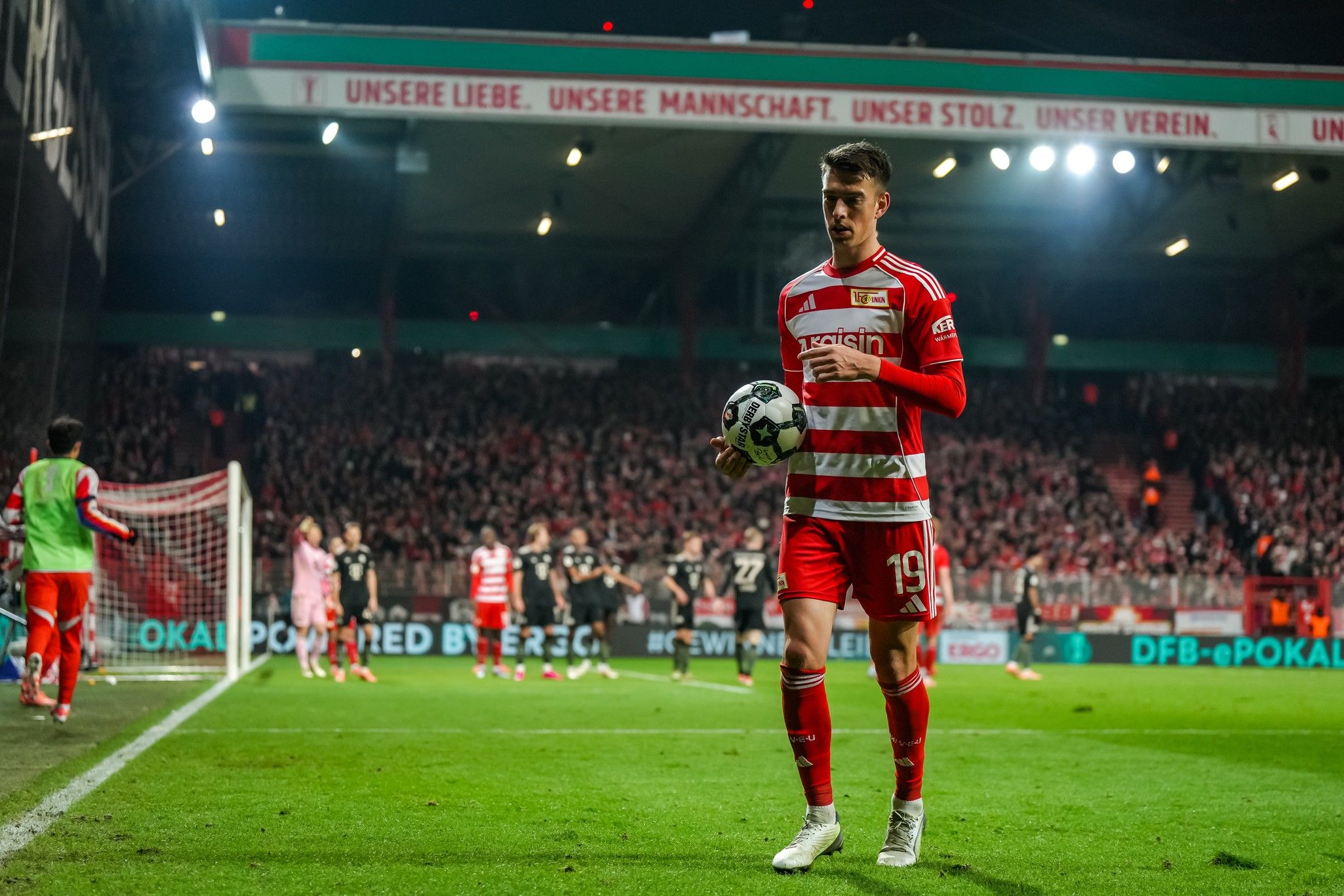 Ein Spieler des FC Union Berlin steht mit einem Ball am Spielfeldrand, während im Hintergrund Fans jubeln.
