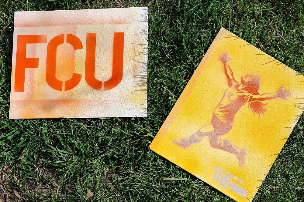 Zwei Sprühbilder auf Rasen: eines mit dem Schriftzug "FCU" in orange, das andere zeigt ein sidendes Kind in gelb.