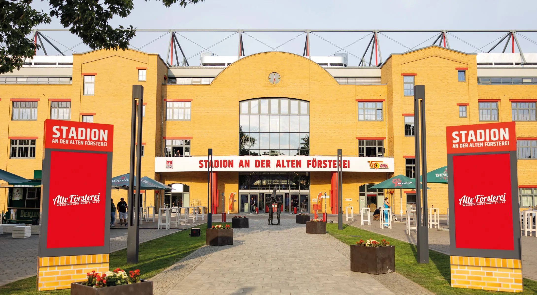 Ein modernes Stadion mit gelber Fassade, Beschilderung und Sitzgelegenheiten im Freien. Bäume und blühende Pflanzen umgeben den Eingangsbereich.