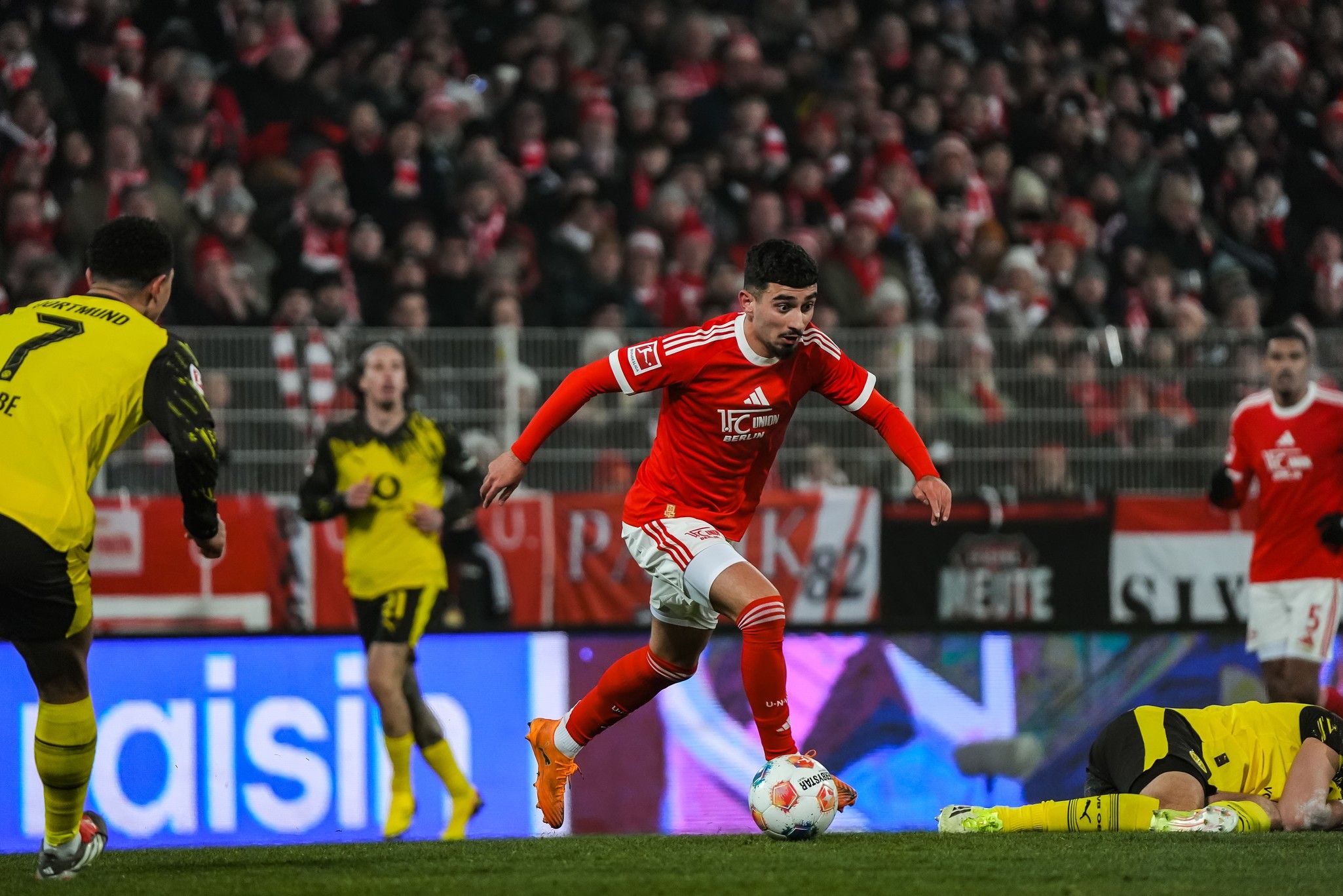 Ein Fußballspieler in rotem Trikot dribbelt den Ball, während andere Spieler in der Nähe stehen. Zuschauer sind im Hintergrund zu sehen.