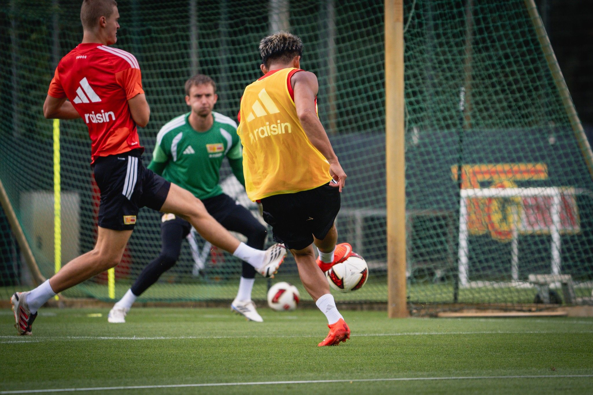 Zwei Fußballspieler in Trainingskleidung spielen auf einem Kunstrasenplatz, während ein Torwart im Hintergrund positioniert ist.
