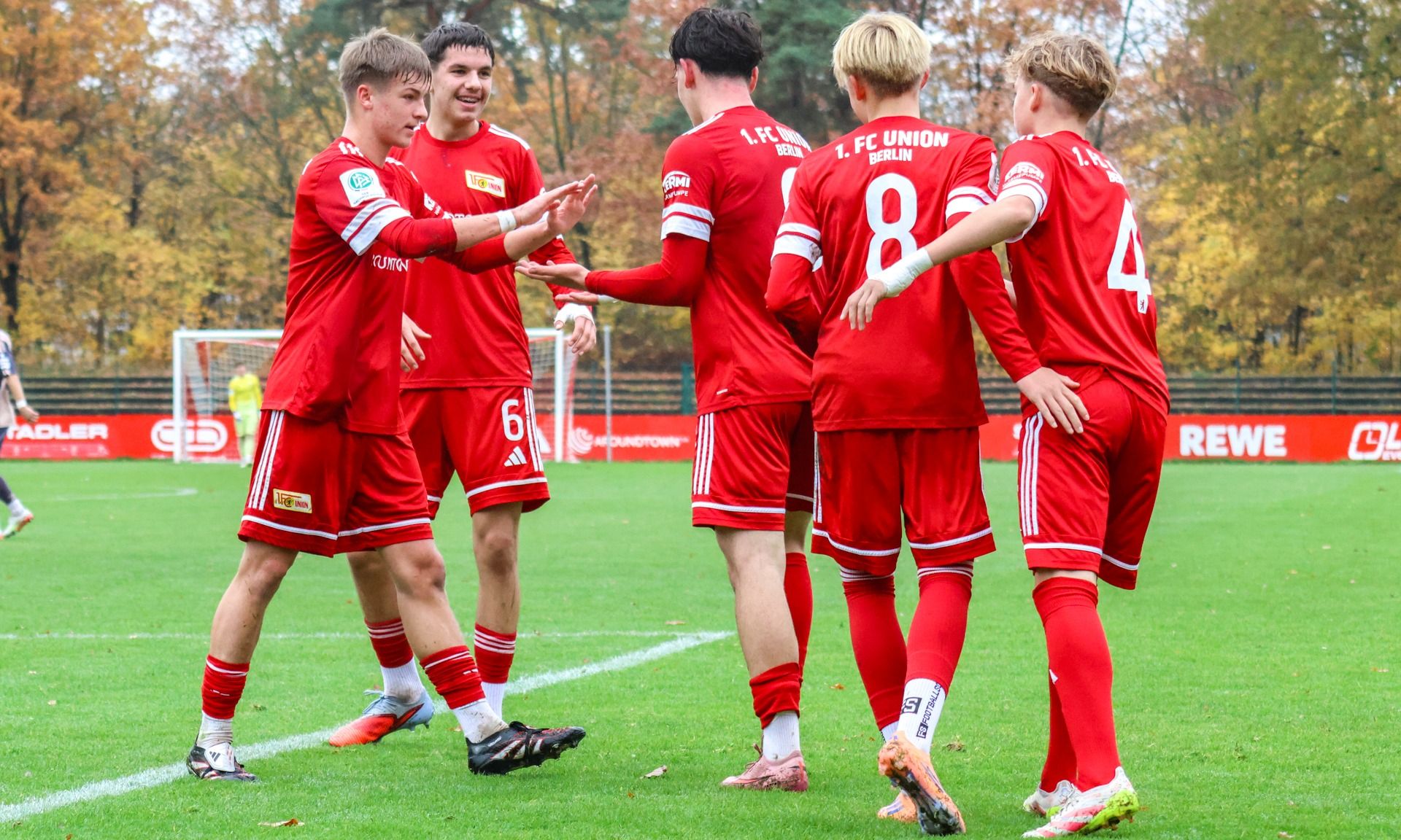 Fünf Fußballspieler in roten Trikots feiern auf dem Platz. Herbstliche Bäume im Hintergrund.