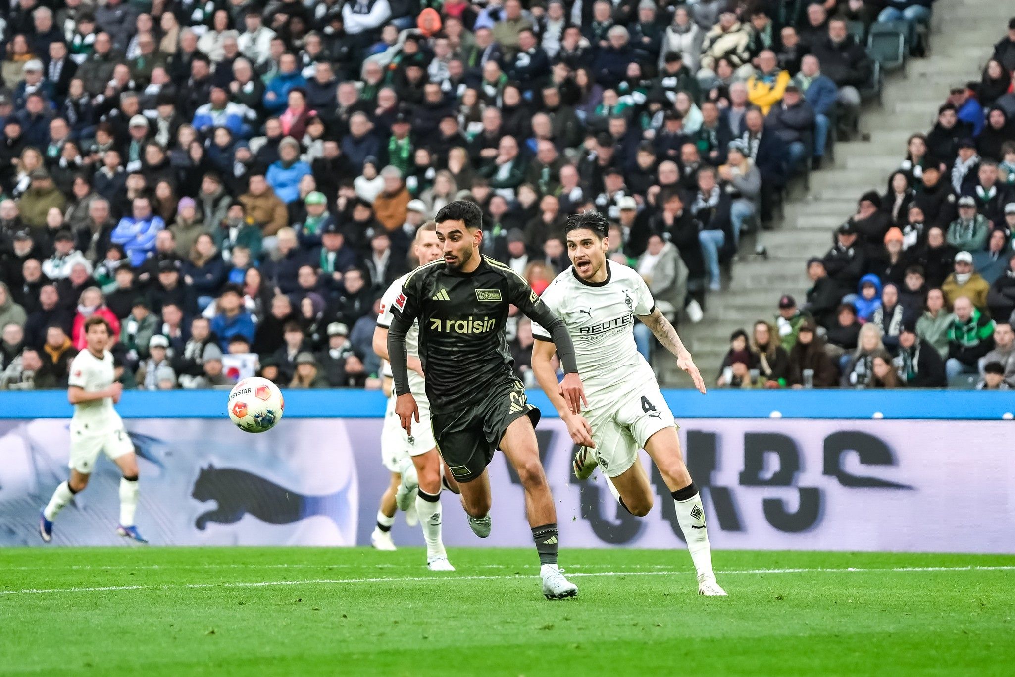 Ein Fußballspieler in schwarzer Kleidung sprintet mit dem Ball, während ein gegnerischer Spieler ihn verfolgt. Menschenmenge im Hintergrund.